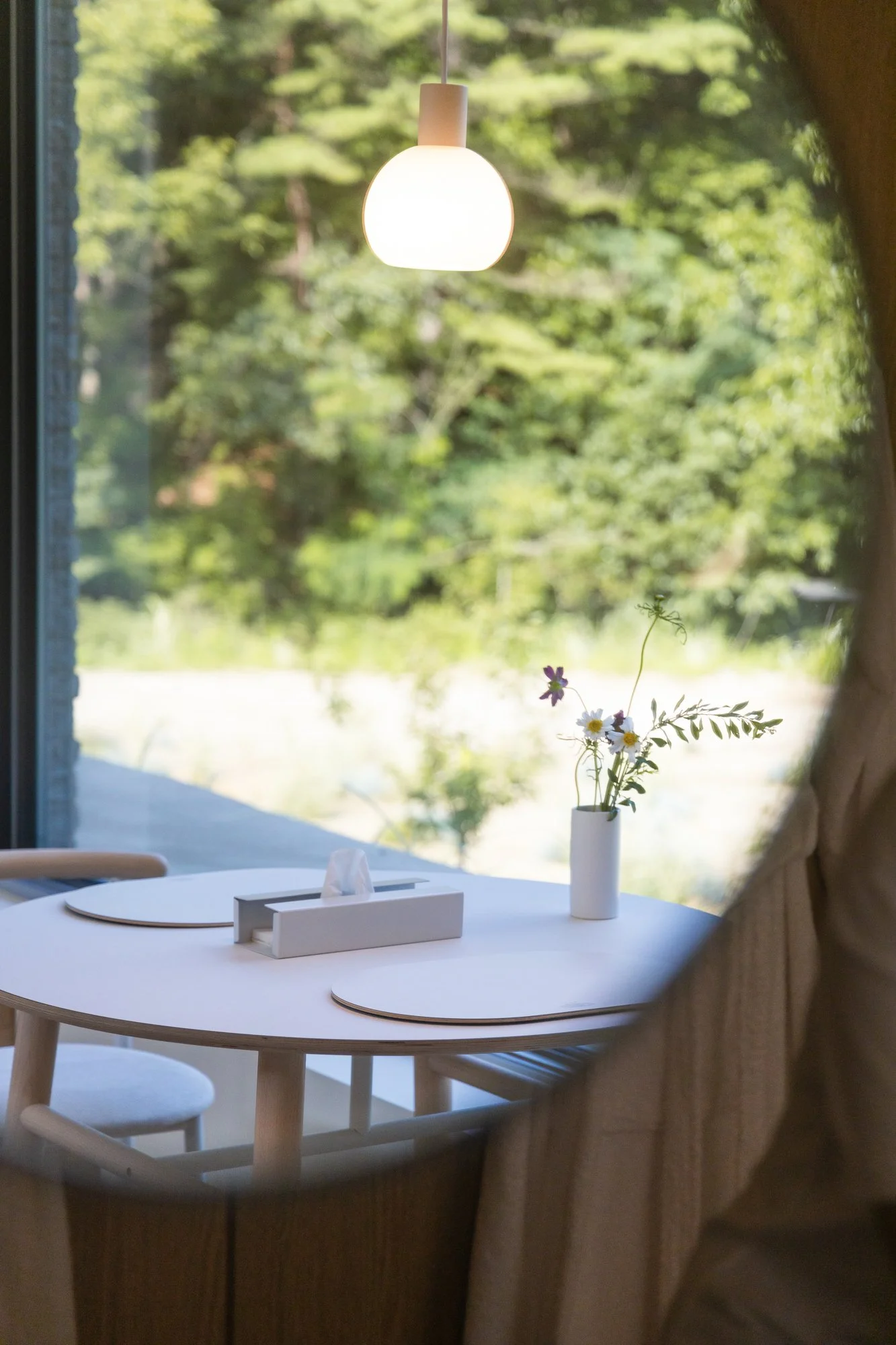 A round white table with a white tissue box and a vase of wildflowers, overlooking a lush green outdoor landscape through large windows with a hanging ceiling light.