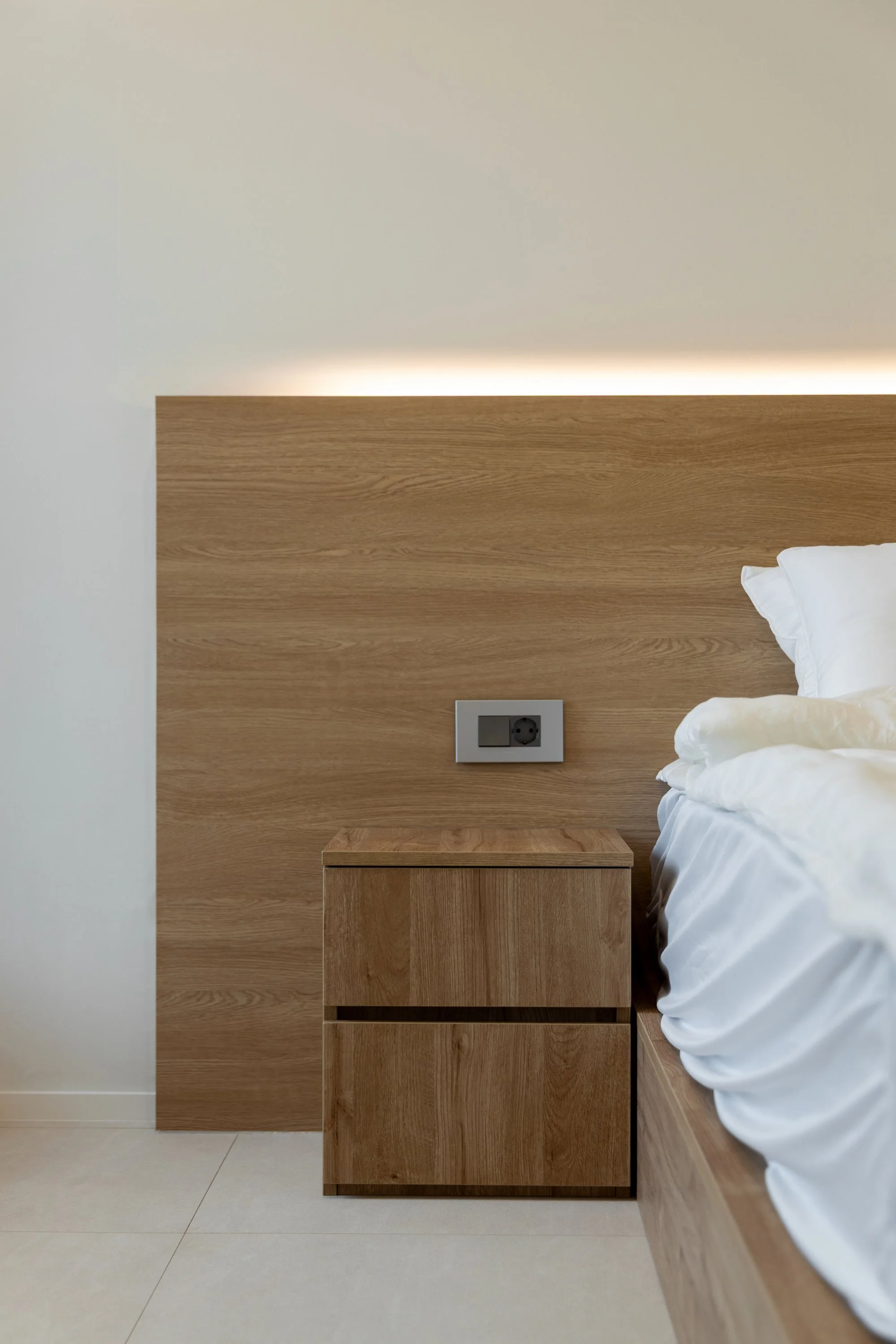 A bedroom with a wooden headboard and bedside table, white bedding, and electrical outlets on the wall.