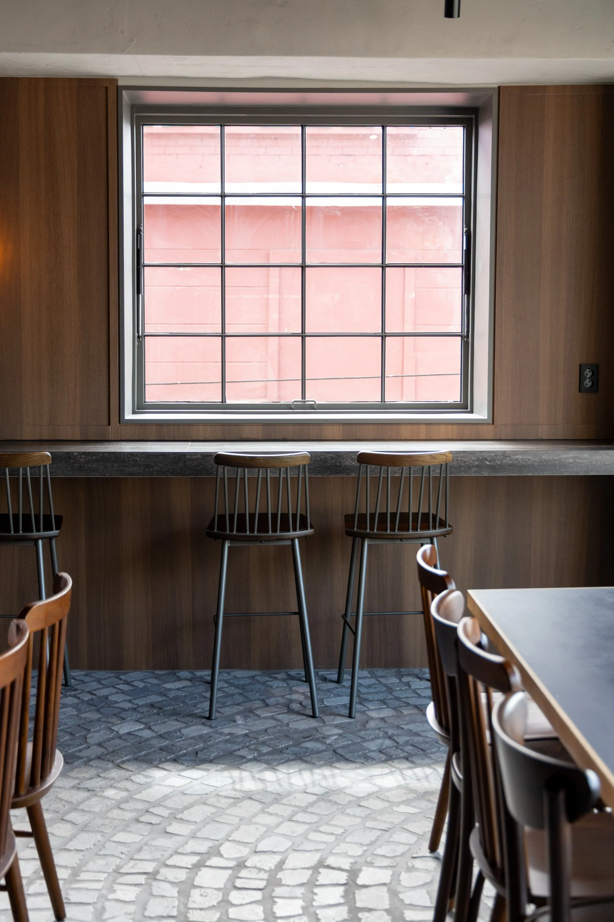 Empty cafe with wooden chairs at a table and a large window with grid pattern, with a view of a red brick wall outside.