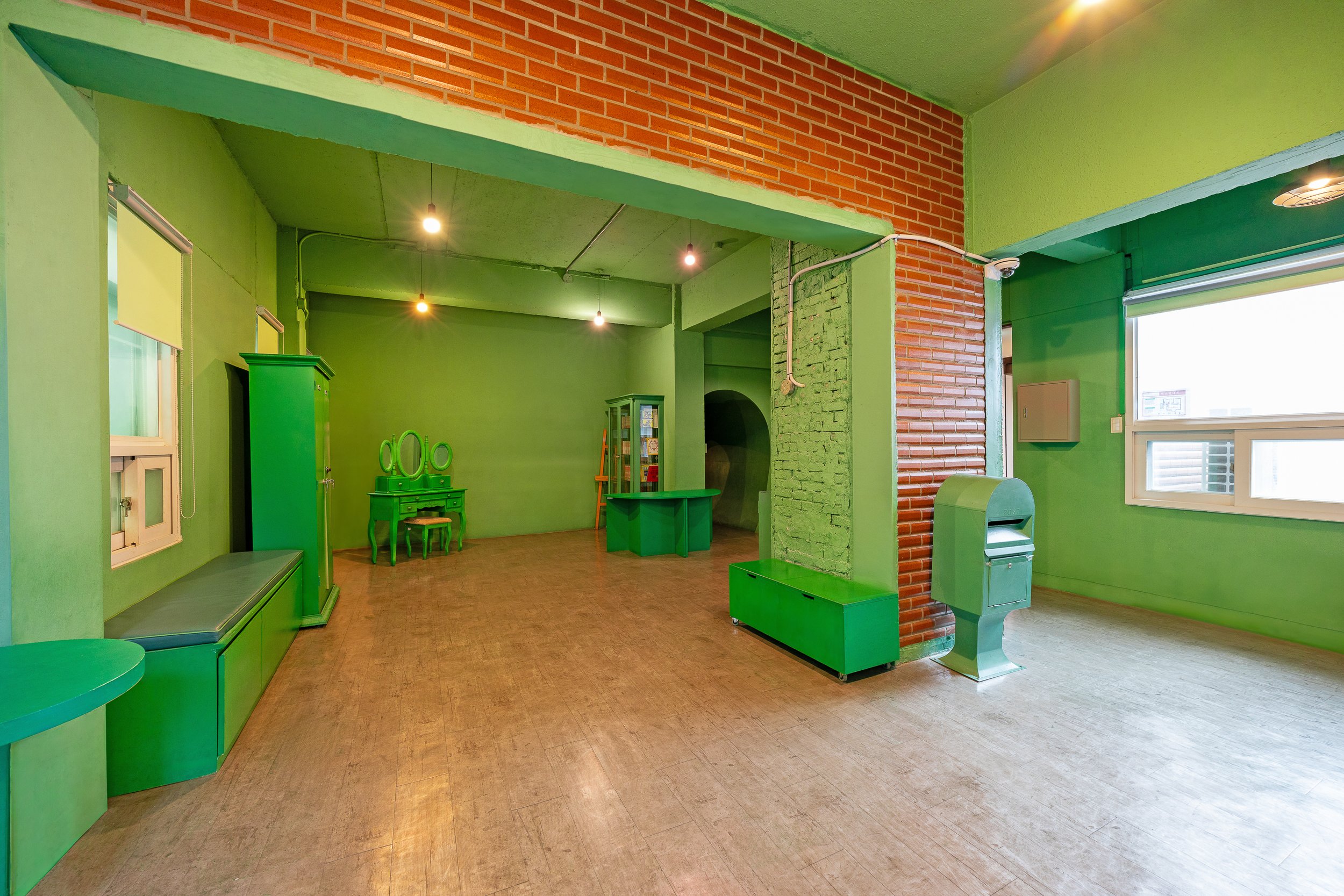 Interior of a room with green walls and furniture, including a bench, wardrobe, vanity with mirrors, and a small green cabinet, with a window on the right and an exposed brick wall.