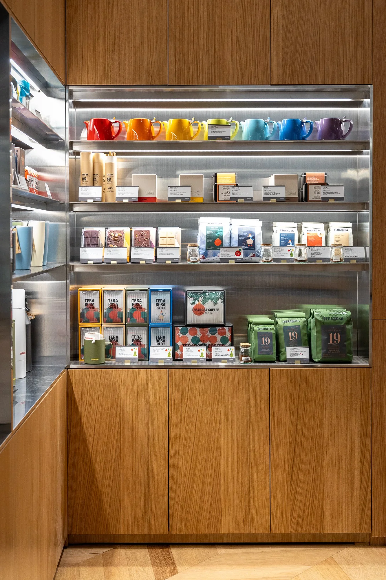 Display shelf with colorful ceramic mugs at the top, various coffee and tea products on the middle and lower shelves, in a wood-paneled section of a store or cafe.