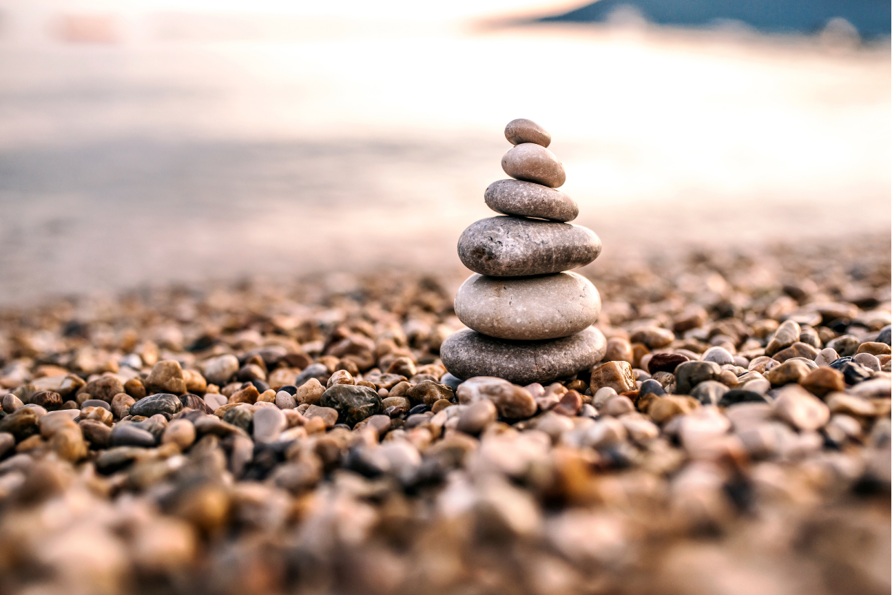 Stack of seven smooth stones placed on a pebbled beach near the water with a blurred shoreline in the background.