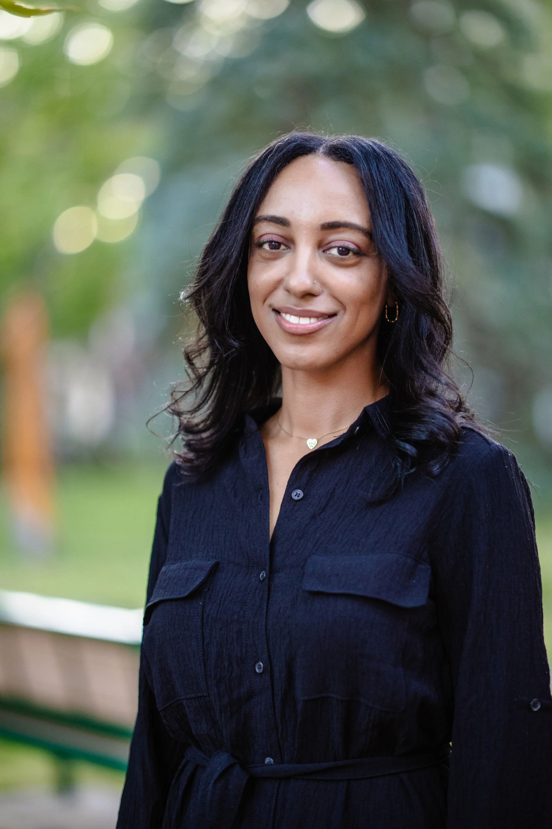 A woman with dark, wavy hair and light brown skin smiling outdoors, wearing a black shirt and gold jewelry.