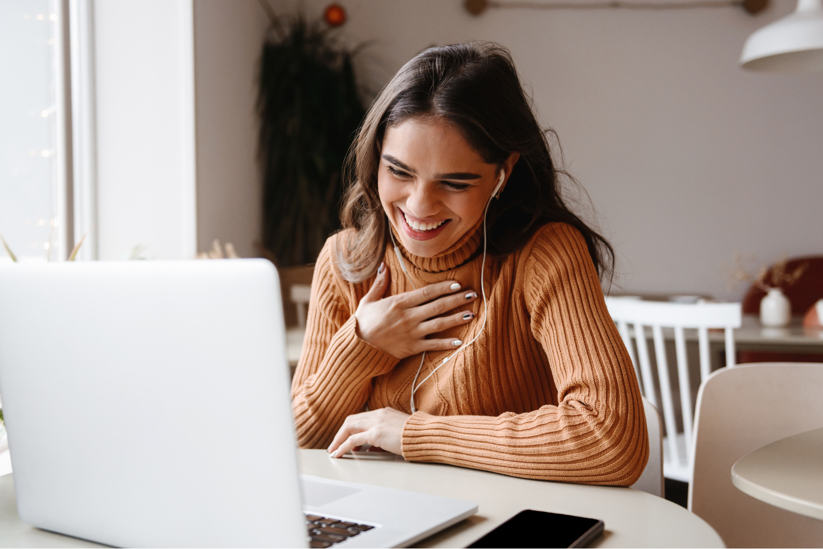 A smiling woman with earphones sitting at a table in front of a laptop, wearing a rust-colored turtleneck sweater, touching her chest with her left hand, in a cozy, well-lit room.