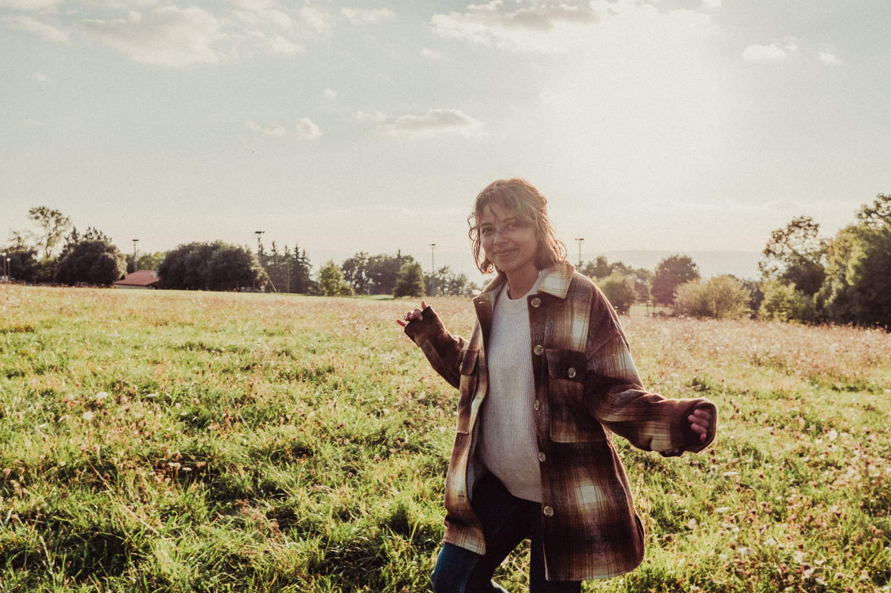 A person with curly hair smiling and standing in a grassy field with trees and a cloudy sky in the background during daytime.