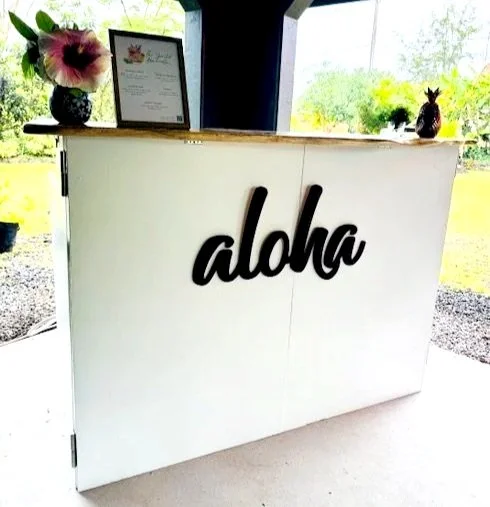 White reception desk with a black 'aloha' sign, decorated with a flower arrangement, framed certificate, and small potted plants inside a building with large windows and outdoor greenery.