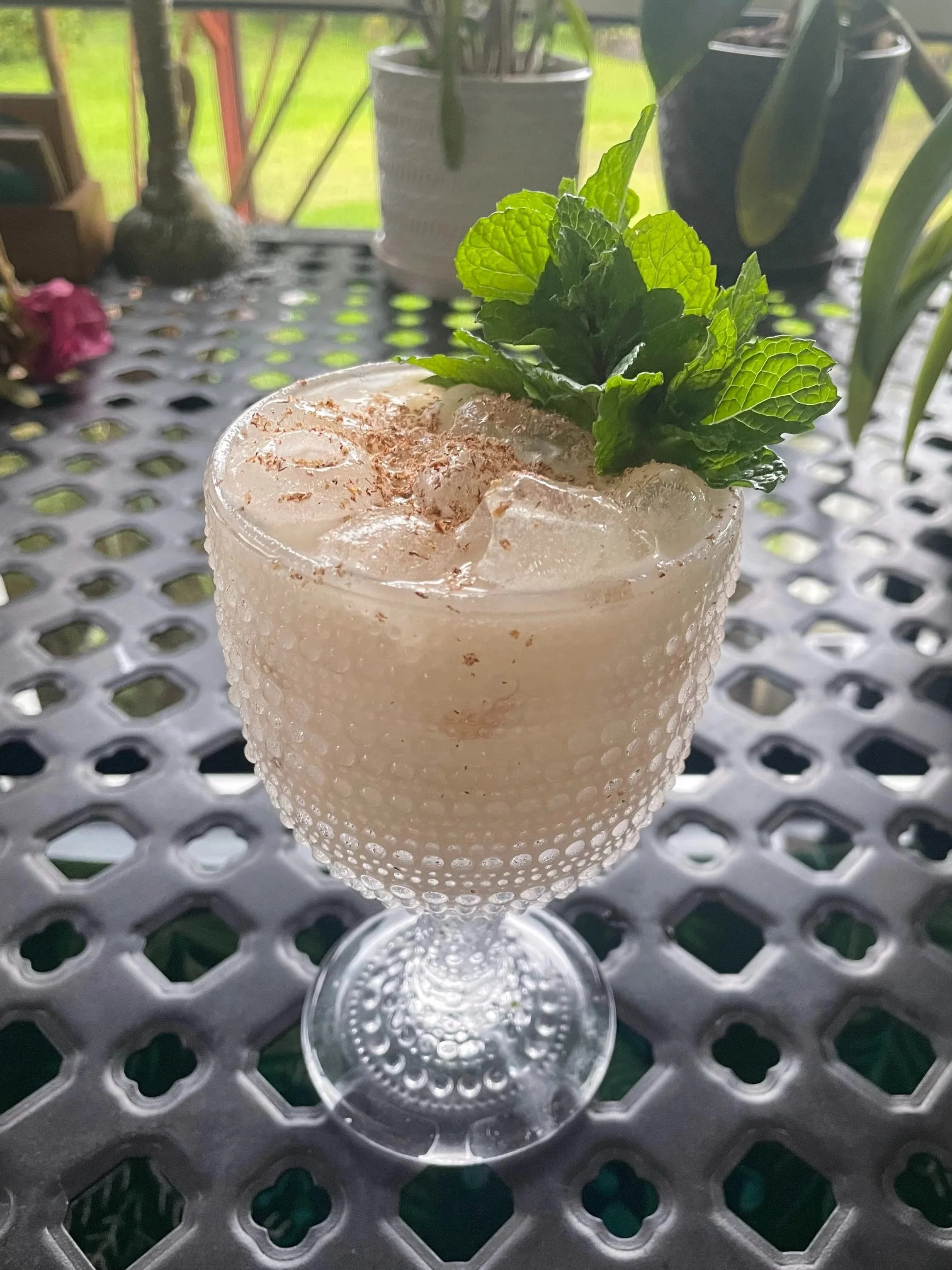 A glass of iced beverage garnished with fresh mint leaves on a perforated metal table, with a potted plant and garden scenery in the background.