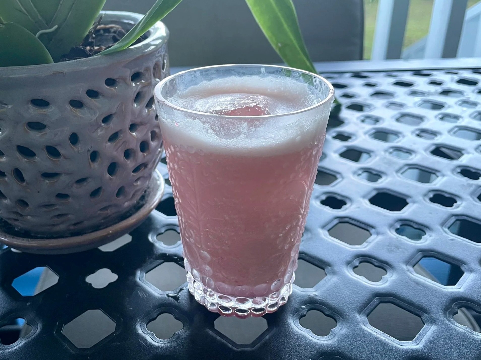 A pink beverage with ice in a textured glass on a black metal table, next to a ceramic pot with a green plant, in an outdoor setting.