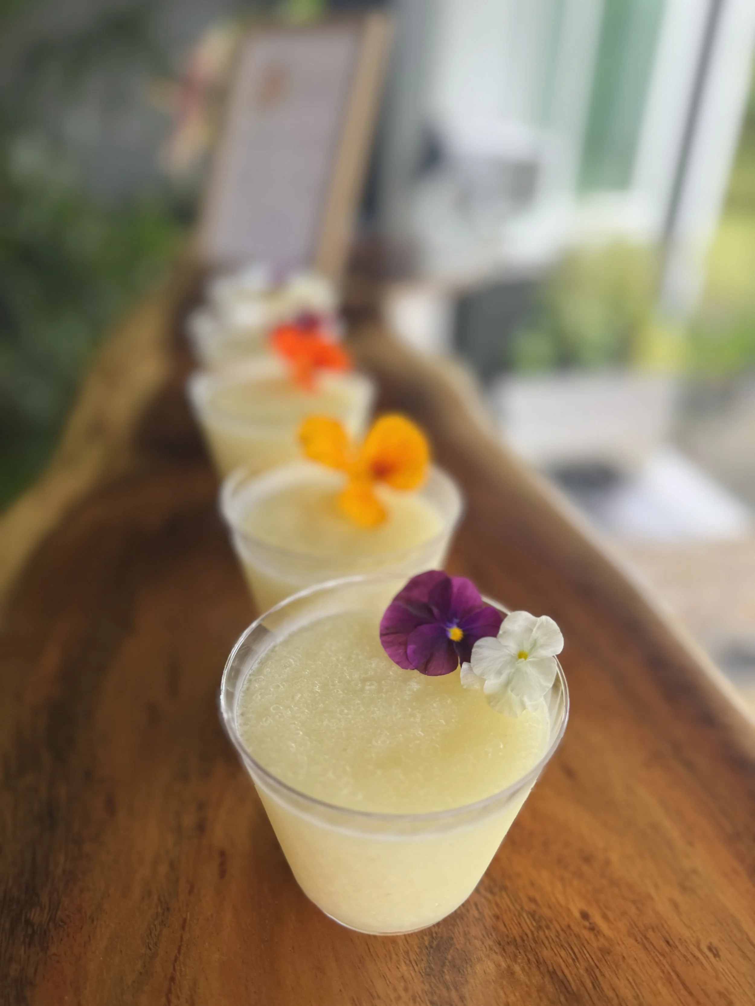 Four cocktails garnished with edible flowers on a wooden surface in front of a blurred window.