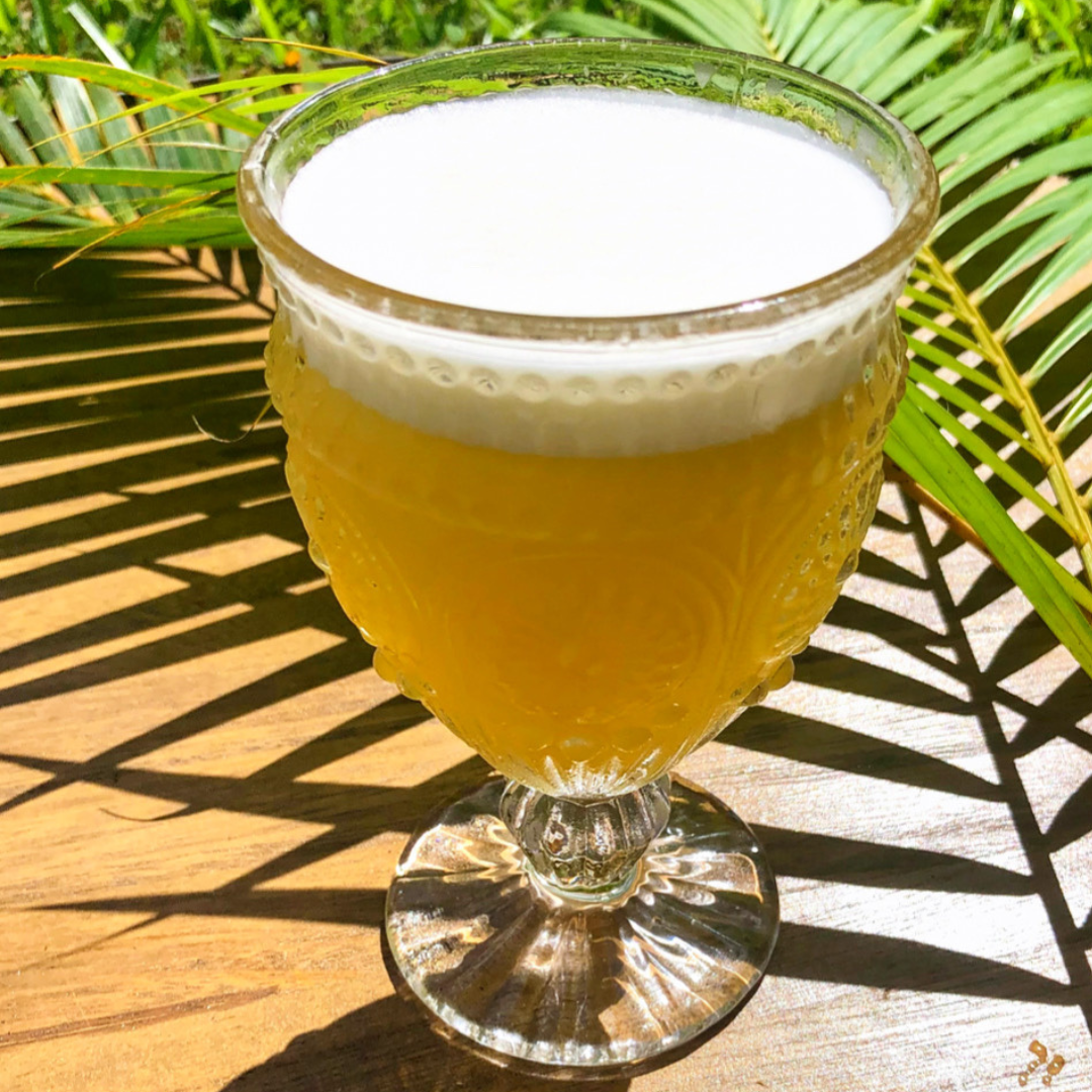 A glass of beer with a frothy head on a wooden table with green palm leaves in the background.