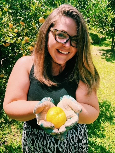 A woman with glasses holding a lemon in her gloved hands in an orchard.