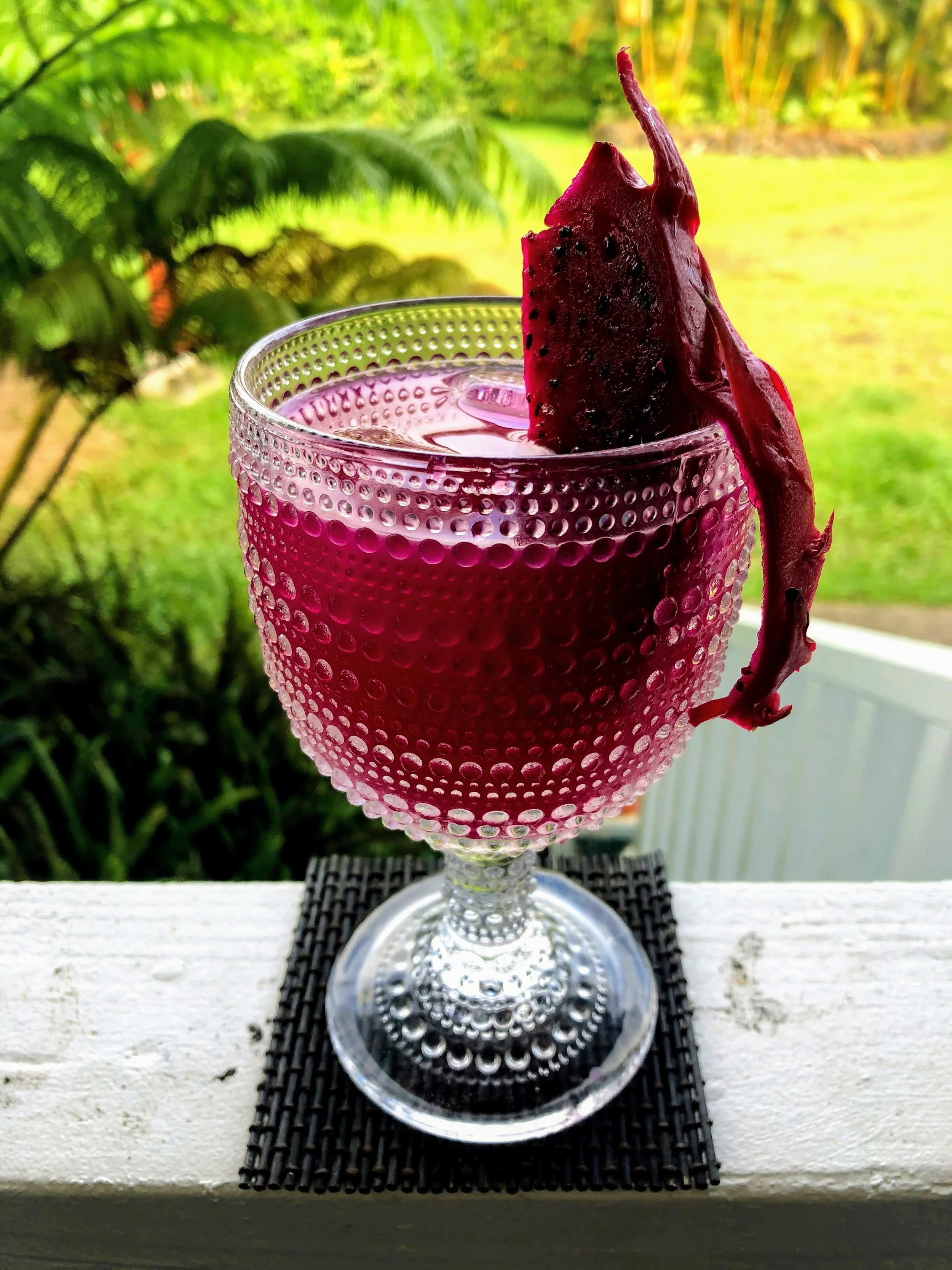 A textured glass with a purple drink and a piece of dragon fruit as garnish, placed on a black mat on a white railing with a garden in the background.