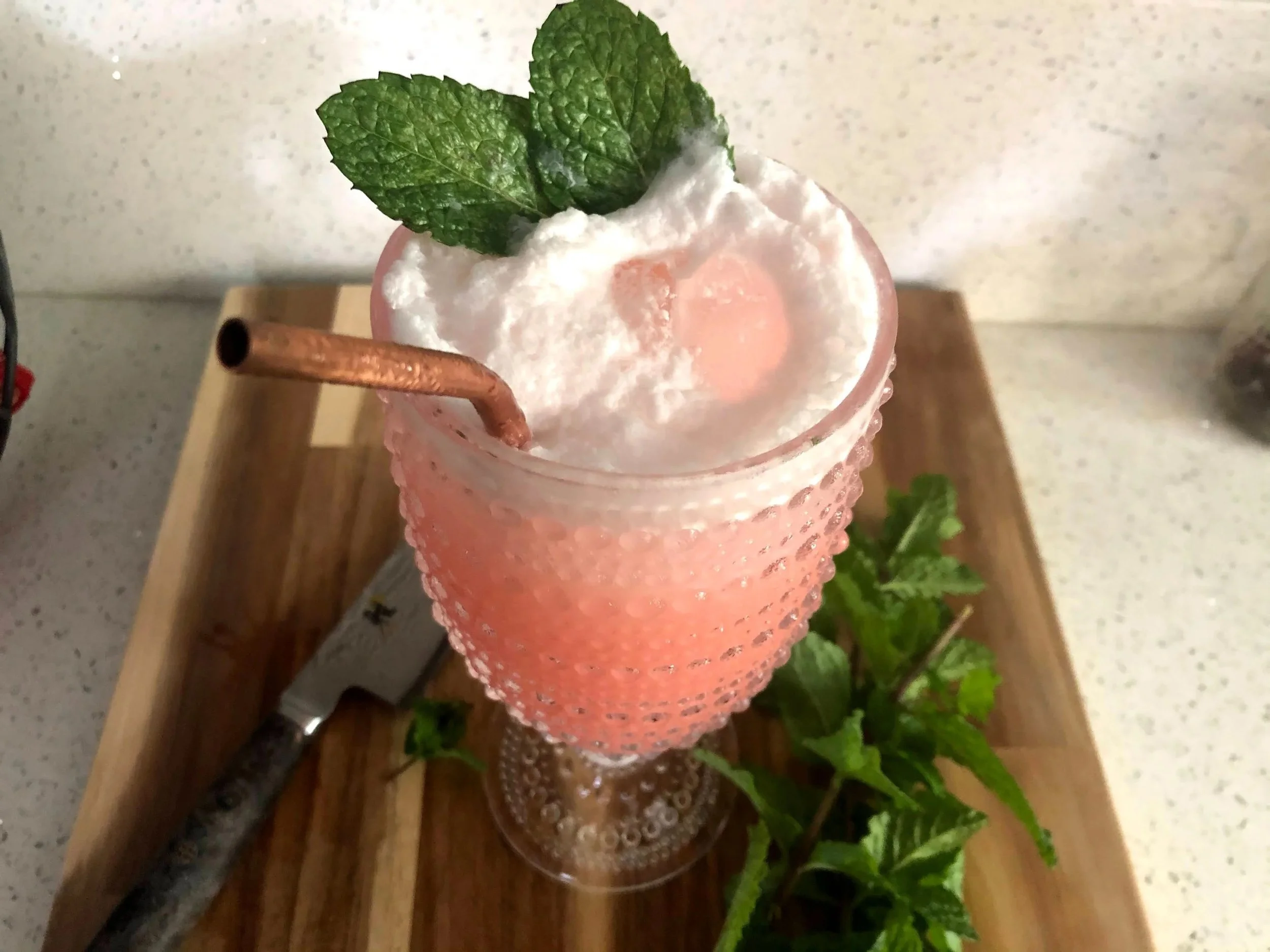 Pink cocktail with whipped cream, fresh mint leaves, a slice of strawberry, a copper straw, on a wooden tray with a sprig of mint and a small knife nearby.