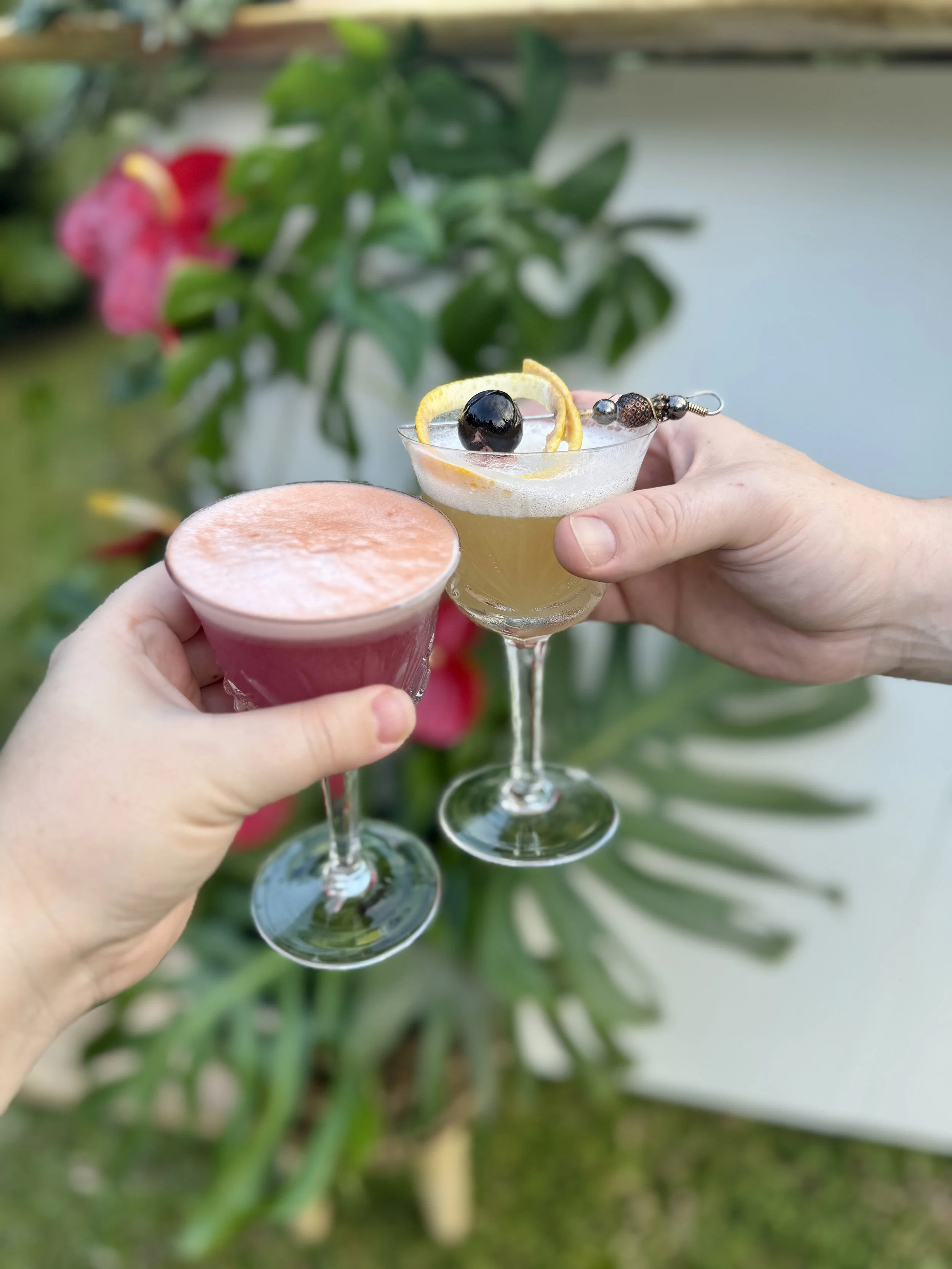 Two hands holding cocktail glasses in an outdoor garden setting, with greenery and pink flowers in the background.