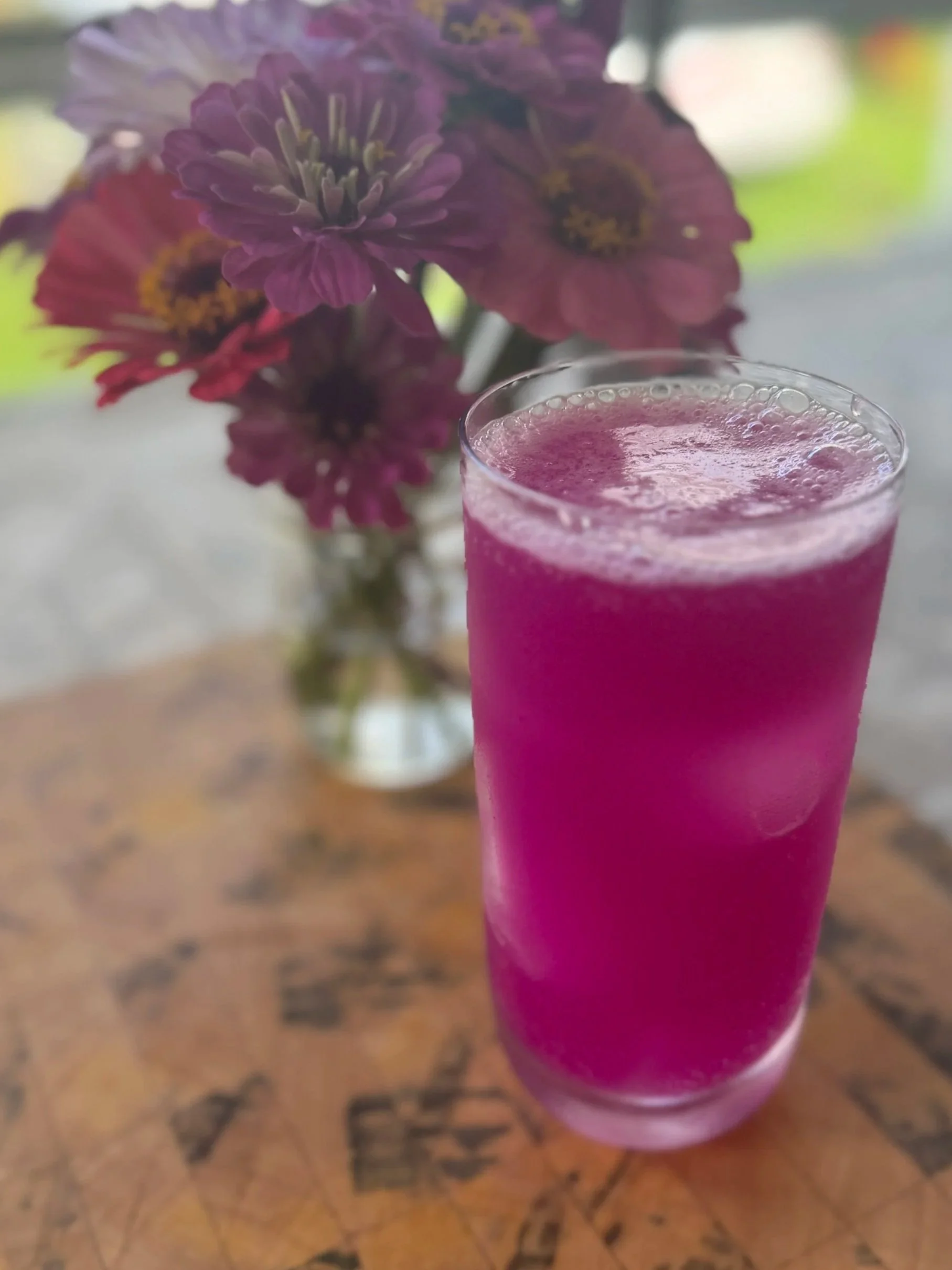 A tall glass filled with a bright pink frozen beverage on a wooden table, with a bouquet of pink and purple flowers in a glass vase in the background.