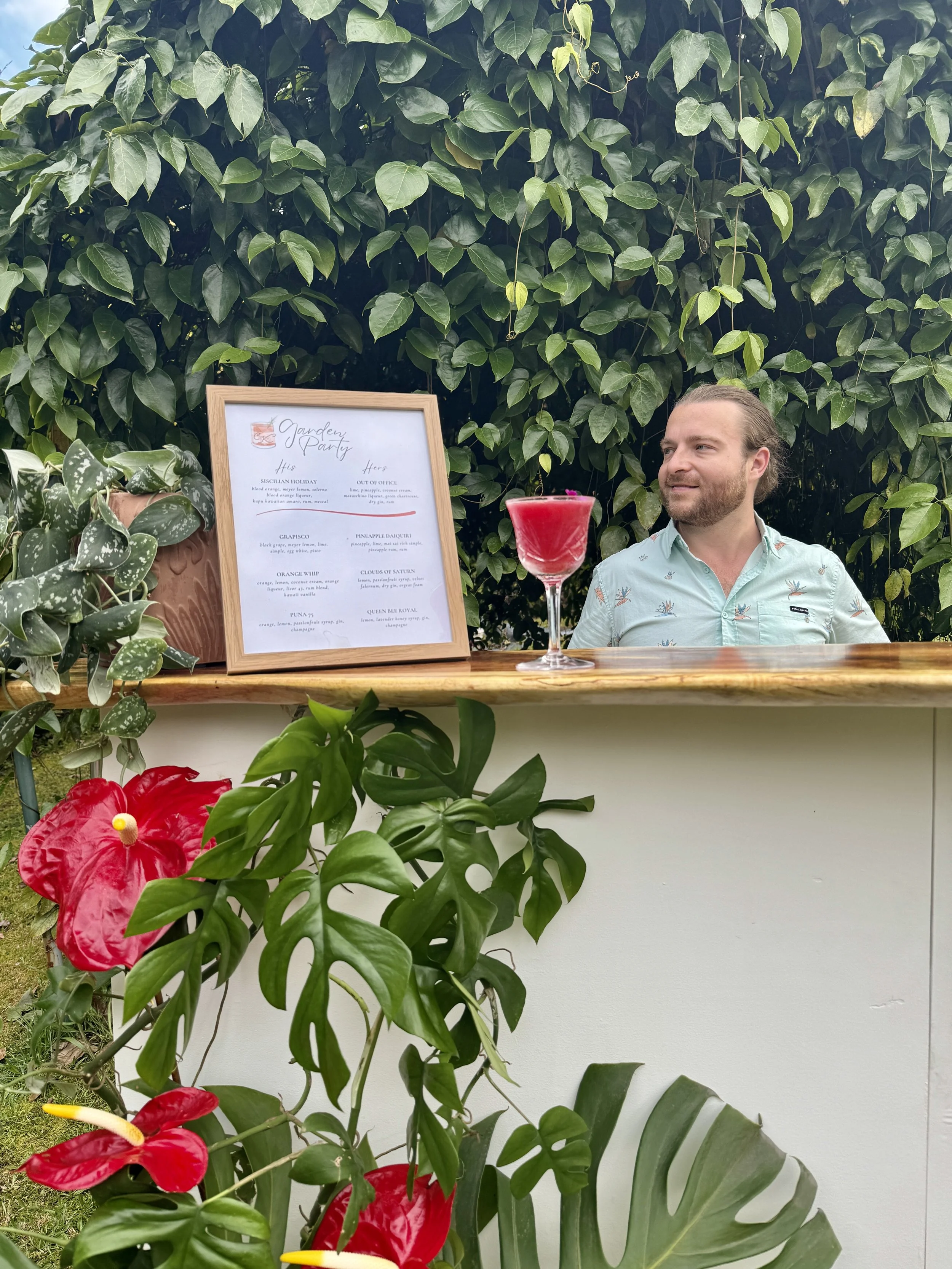 Kirk behind a bar with tropical plants and a framed menu, surrounded by green foliage and vibrant flowers.