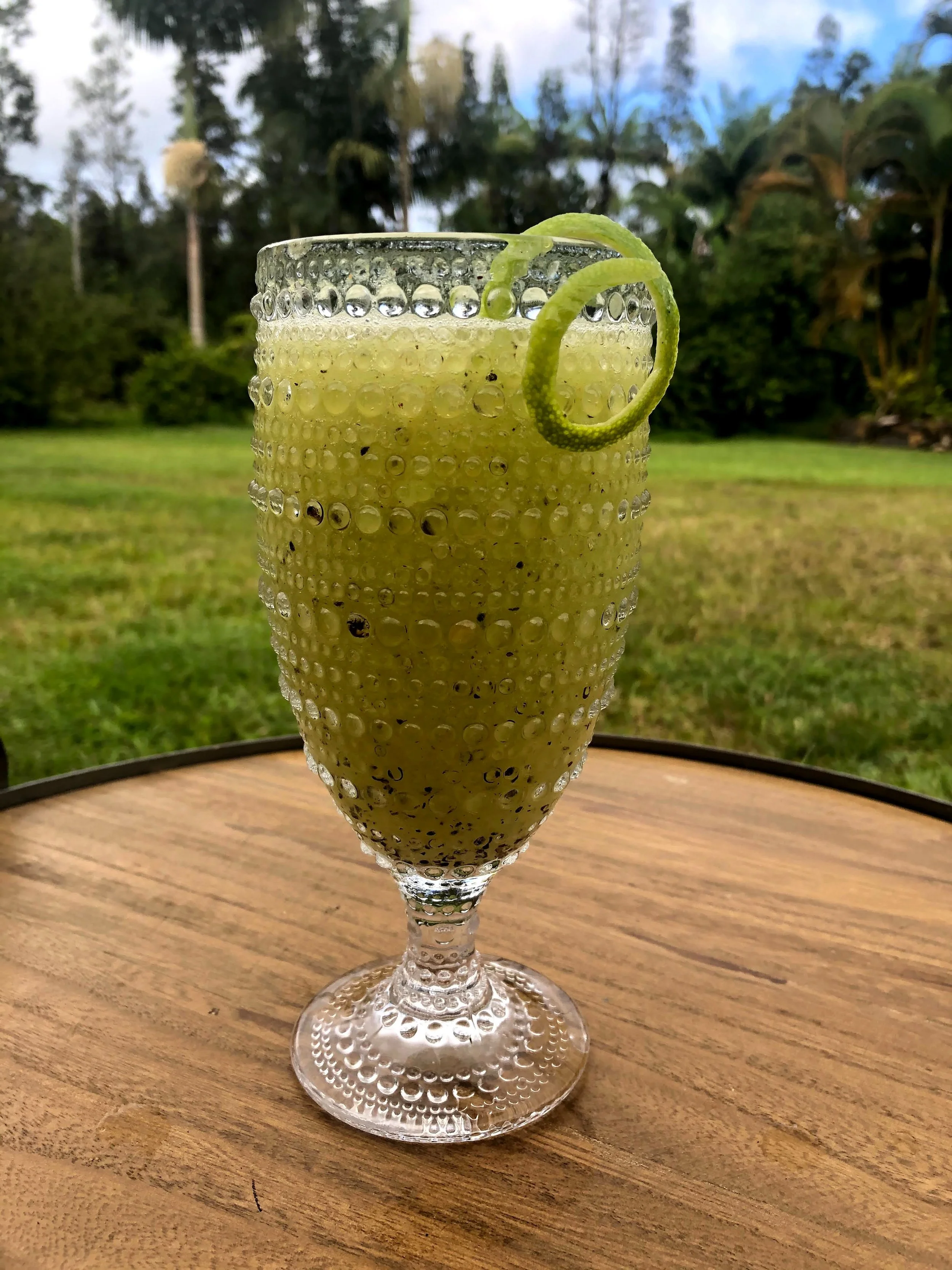 A tall, textured glass filled with a green smoothie, garnished with a peel of citrus twisted into a loop, placed on a wooden table outdoors with a grassy area and trees in the background.