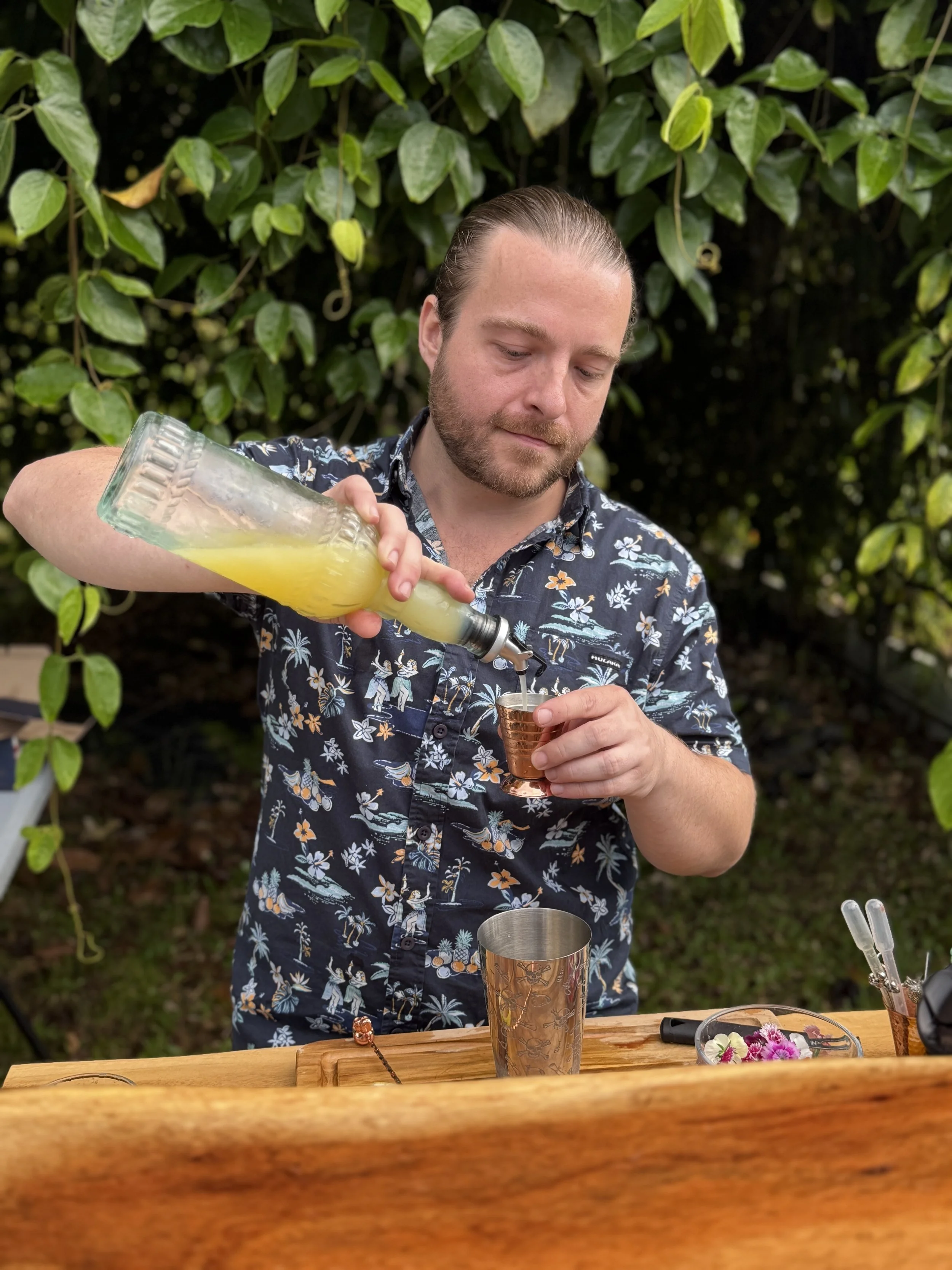 A man with a beard and long hair tied back, wearing a Hawaiian shirt, pours a yellow beverage from a glass bottle into a copper cup at an outdoor bar or tiki setup with greenery in the background.