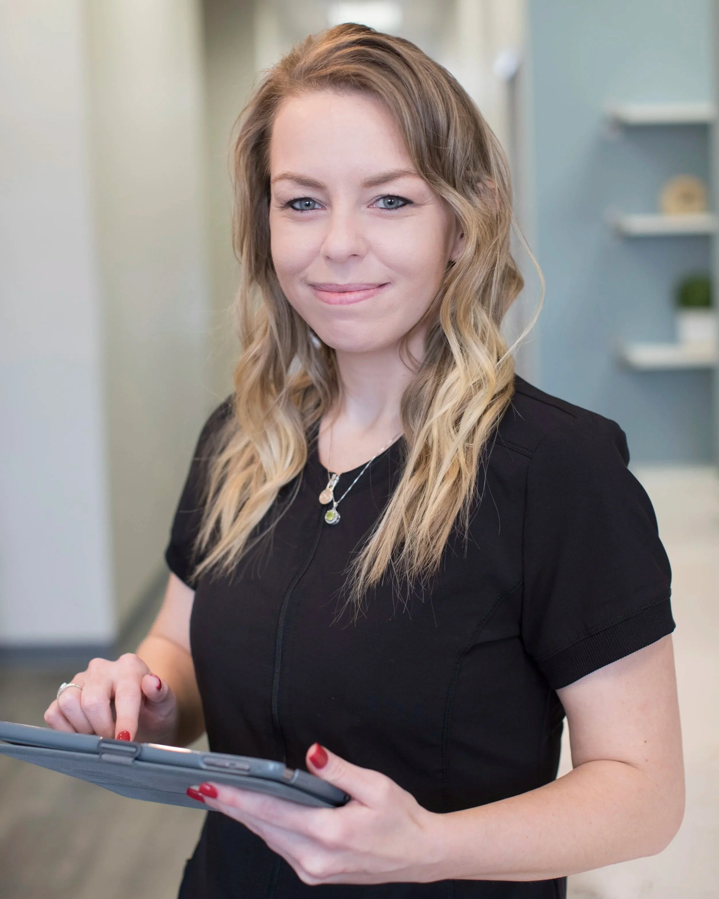 A woman with long wavy blonde hair, wearing a black top, holding a tablet device while looking at it and smiling. She has red nail polish and wears a silver necklace, in an indoor setting with blurred shelves in the background.
