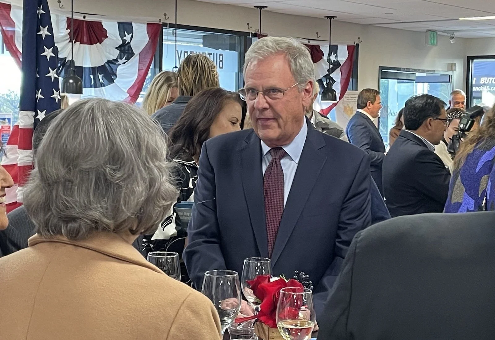 A man in a dark suit and red tie talking to an older woman with gray hair, seated at a table with wine glasses and a small bouquet of red flowers. People are mingling in the background with patriotic decorations and American flags.