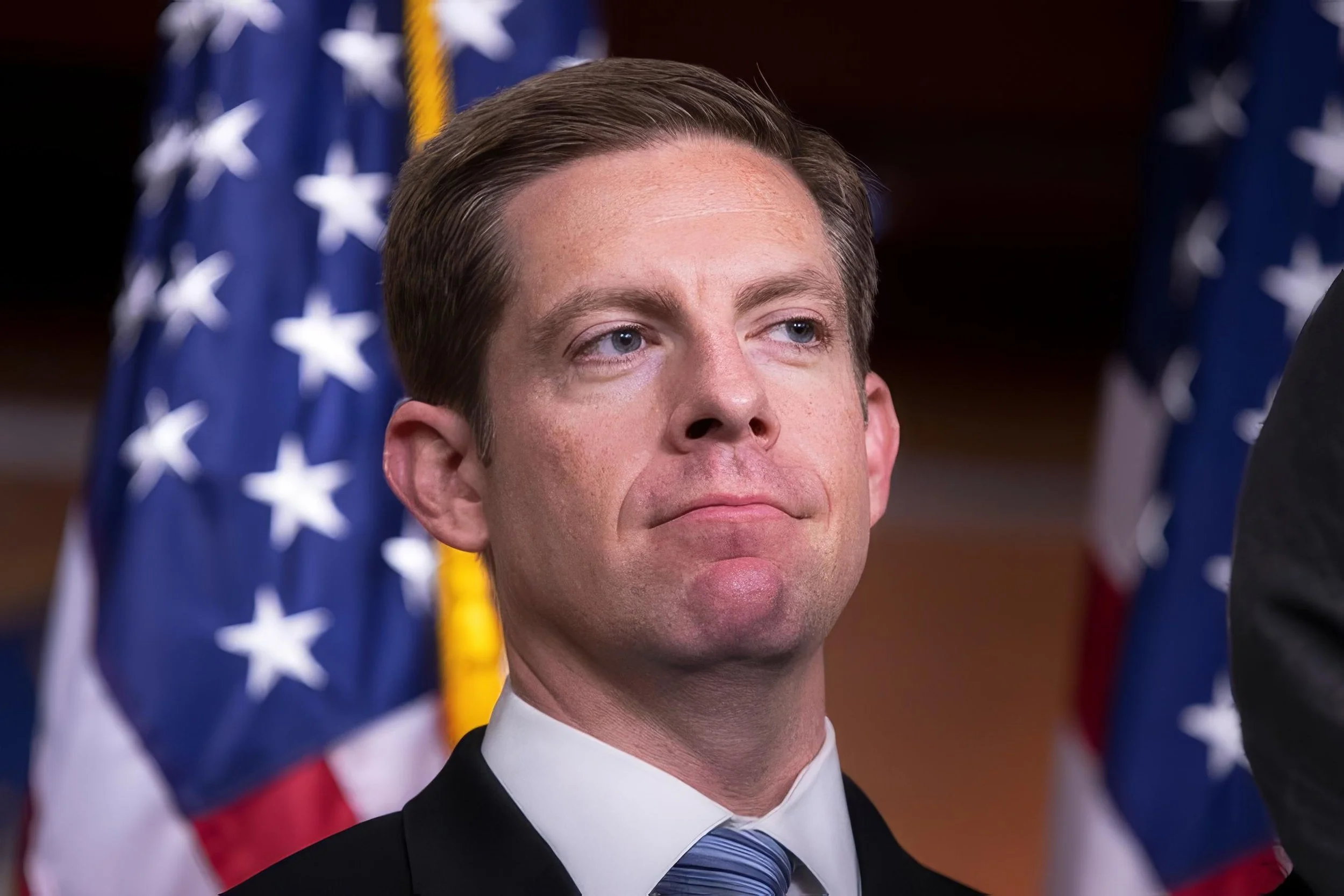 A man in formal suit with a striped tie standing in front of American flags.