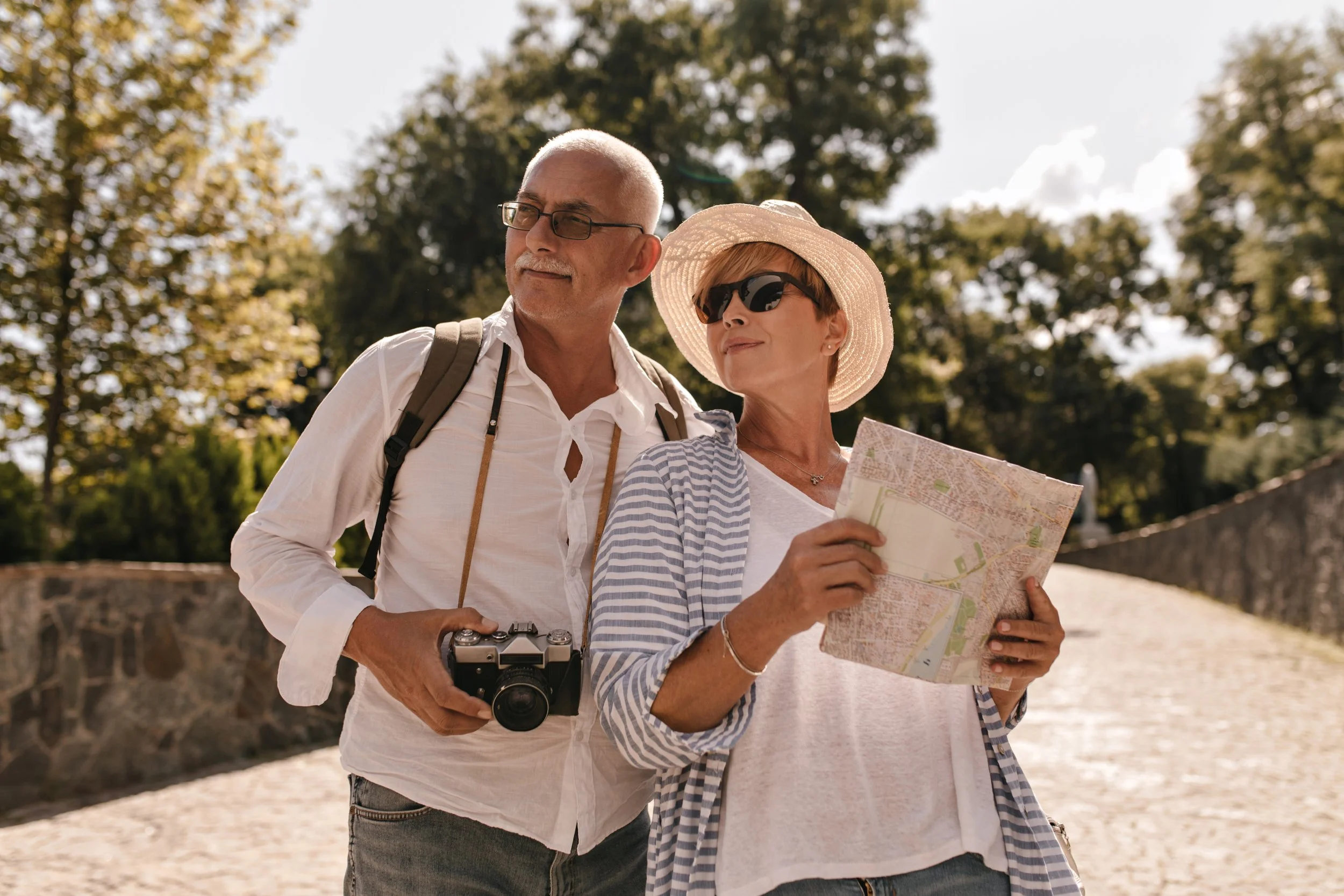 An older man and woman are walking outdoors in a park, with the woman holding a map and the man carrying a camera. They are dressed casually, with the woman wearing a sun hat and sunglasses, and the man wearing glasses with a backpack.