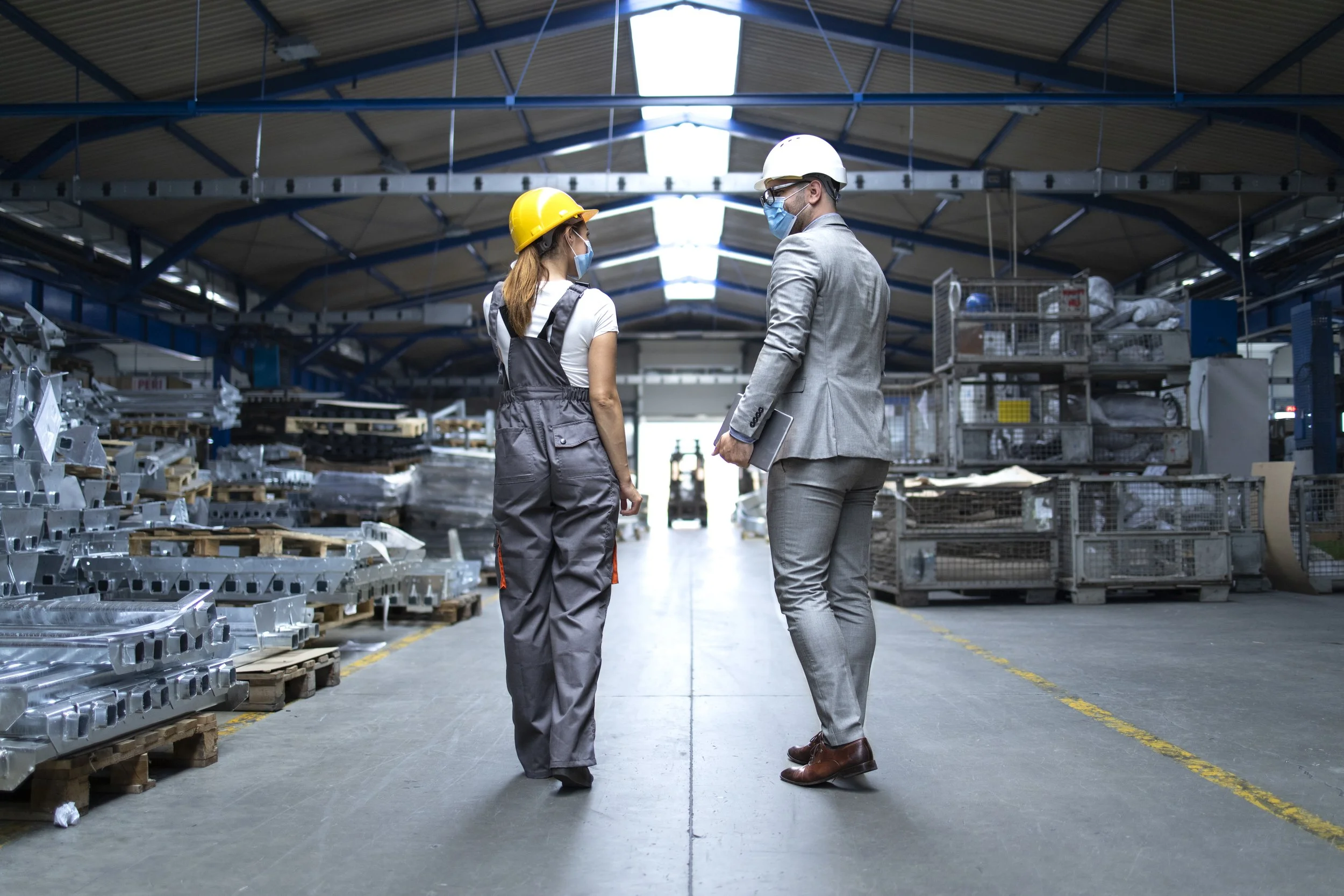 Two people in a warehouse wearing safety helmets and masks, having a conversation. One appears to be a woman in gray work overalls with a yellow helmet, the other a man in a gray suit with a white helmet, holding a tablet.