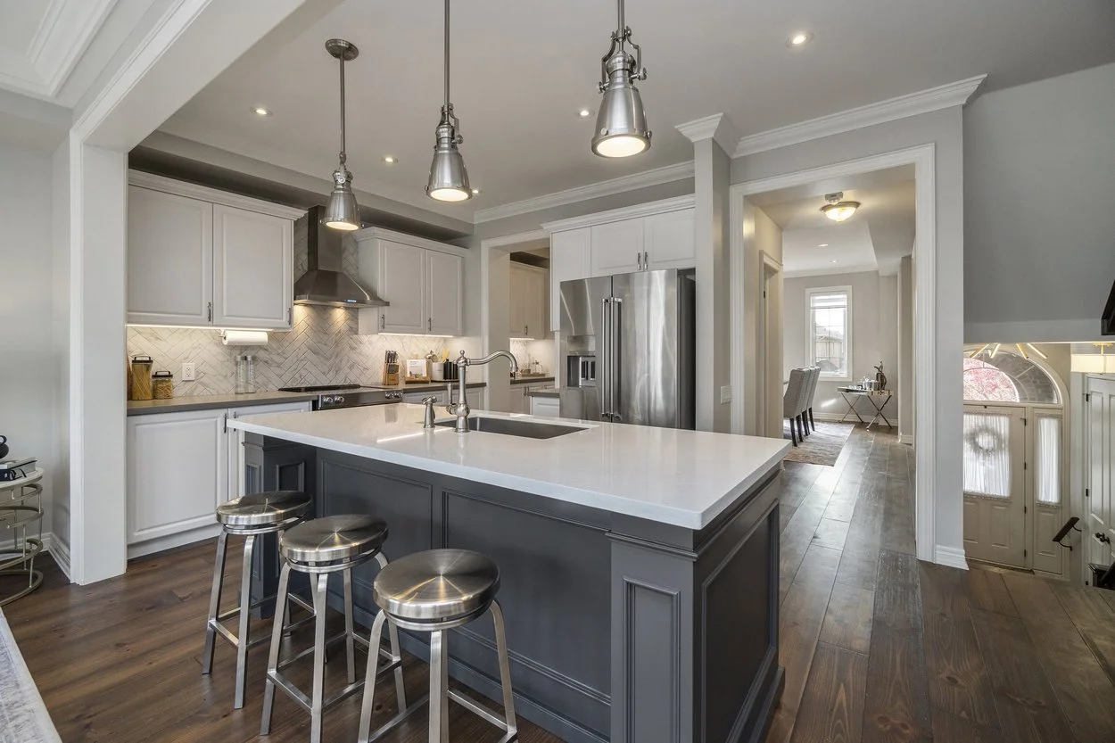 Modern kitchen with a gray island and pendant lights, stainless steel refrigerator, white cabinetry, and a view into a dining area.