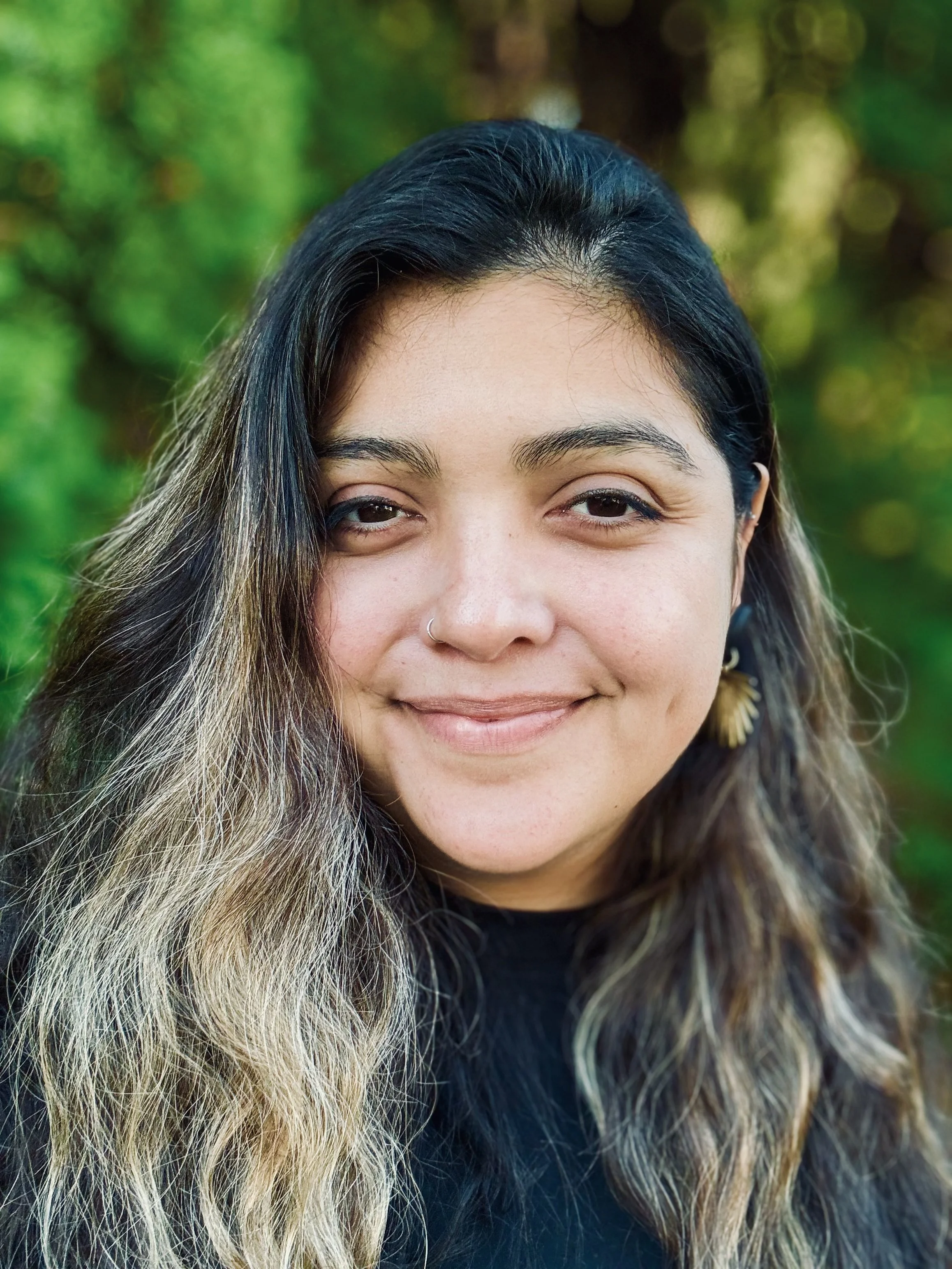 A young woman with long, wavy hair, wearing a nose ring and earrings, smiling outdoors with a background of greenery.