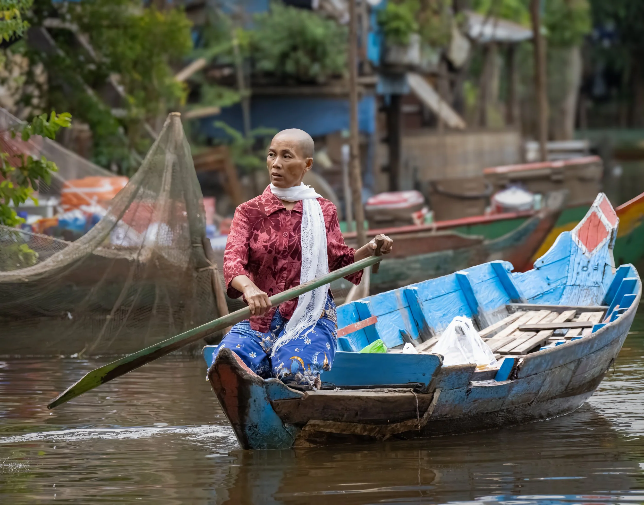 A woman in a red blouse and blue skirt paddles a blue wooden boat on a river, with several other boats and a stilt village in the background.