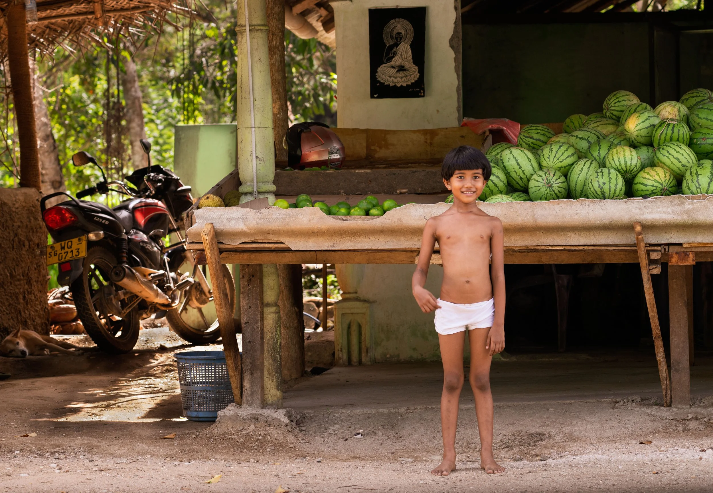 This picture captures the almost universal food stand in front of most homes in rural Sri Lanka. The motorcycle is the family's mode of transportation, often with 3-4 people on board. The adorable Sri Lankan child, standing proudly in white underwear
