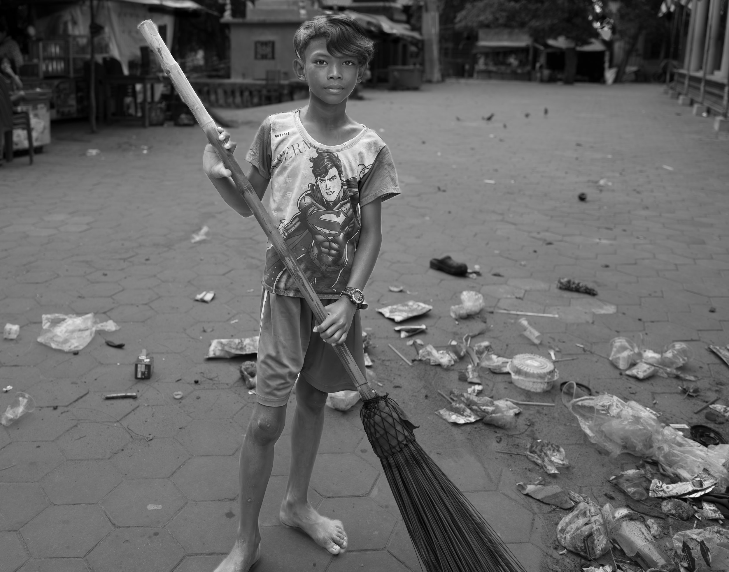 A young boy wearing a Superman t-shirt and shorts is barefoot, holding a broom and standing on an empty street. The street is littered with trash and debris.