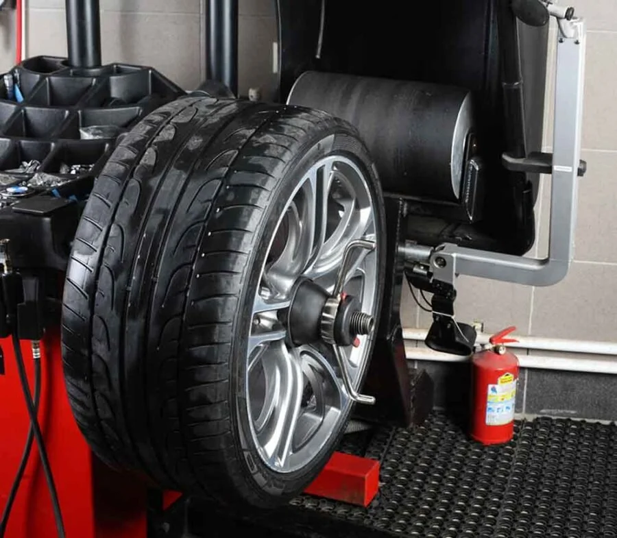 A tire mounted on a wheel with a vibration balance machine in a garage or workshop, with a fire extinguisher nearby.