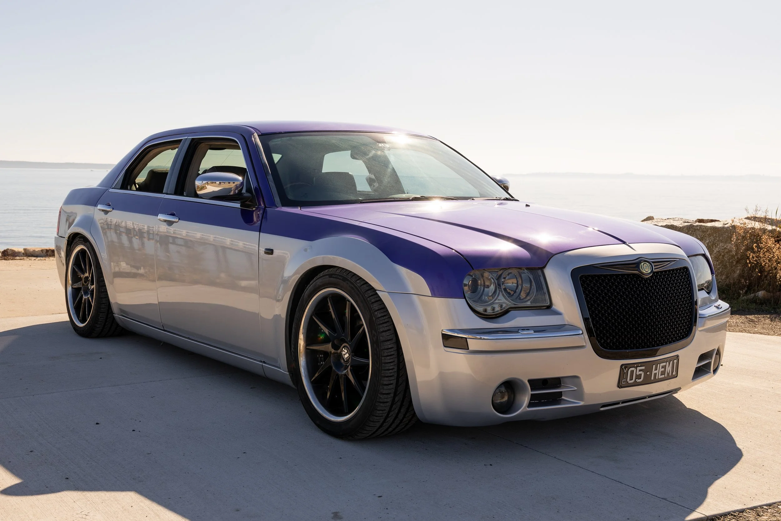 A stylized black and silver sedan parked on a concrete surface near the water, with a large front grille, tinted headlights, and custom wheels, under a clear sky.