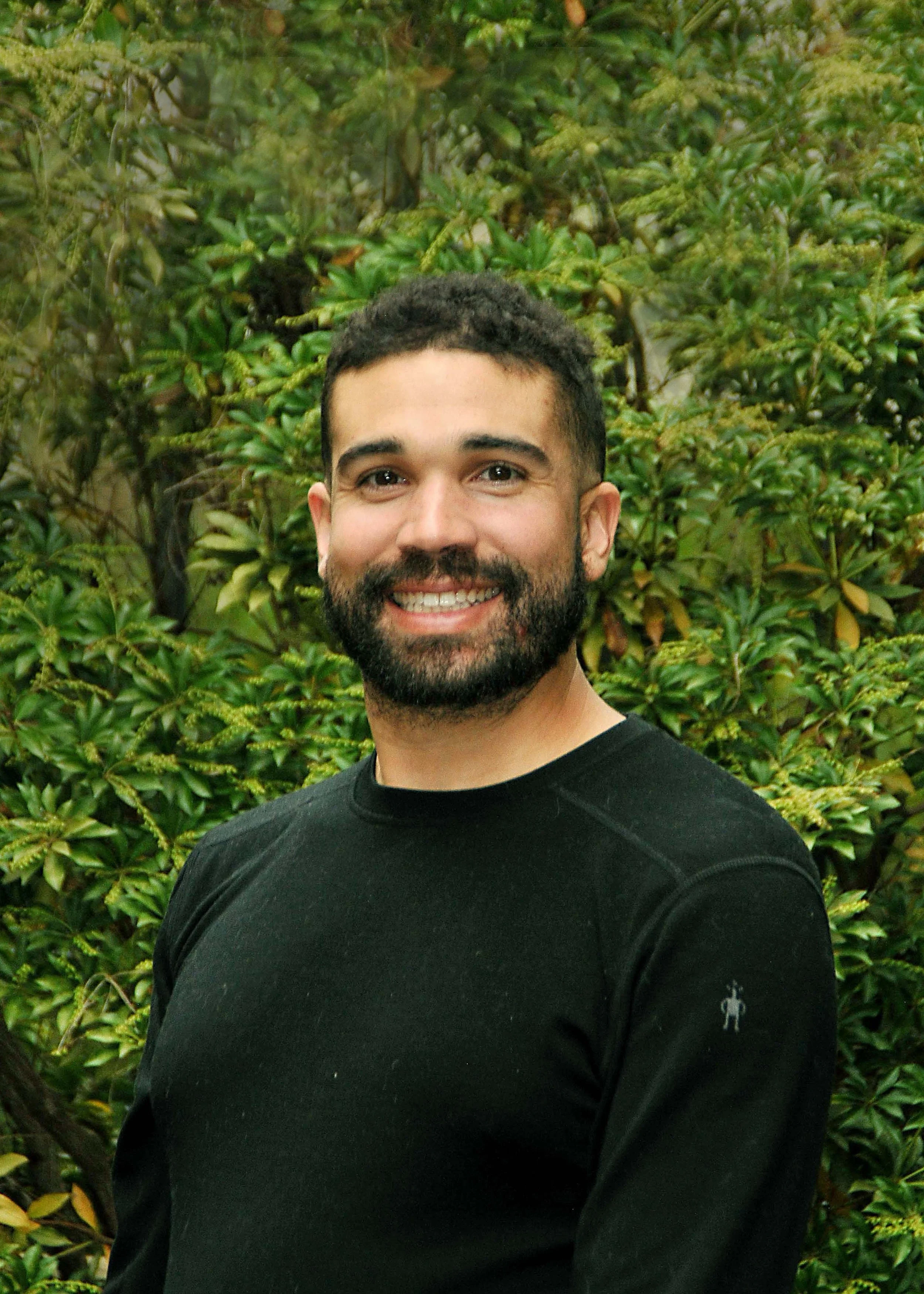 A smiling man with dark curly hair and a beard, wearing a black long-sleeve shirt, standing outside in front of lush green bushes.