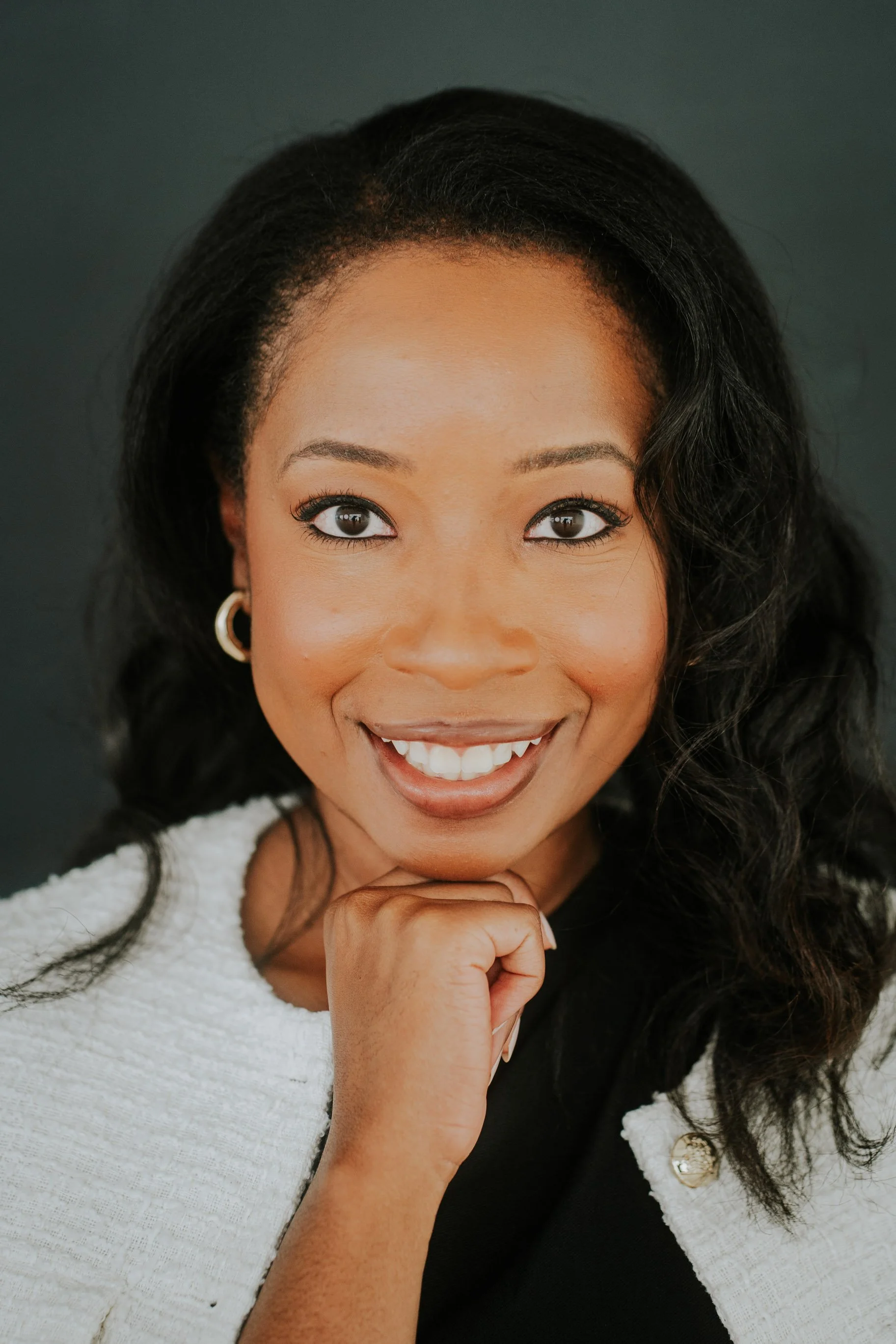 A smiling woman with curly black hair, wearing pearl earrings, a white textured jacket, and a black top.
