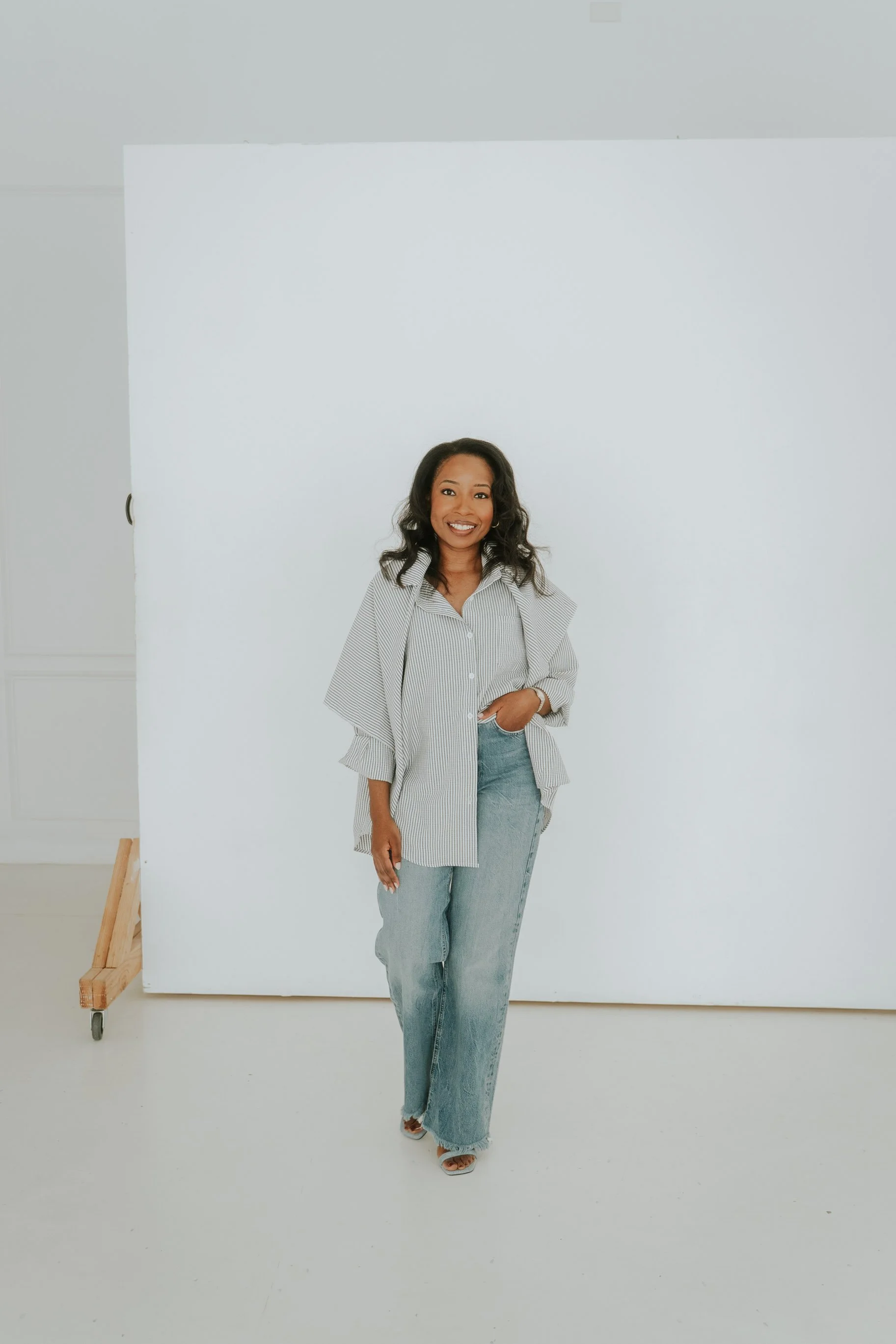 A woman smiling and posing in front of a white backdrop, dressed in a striped shirt and jeans.