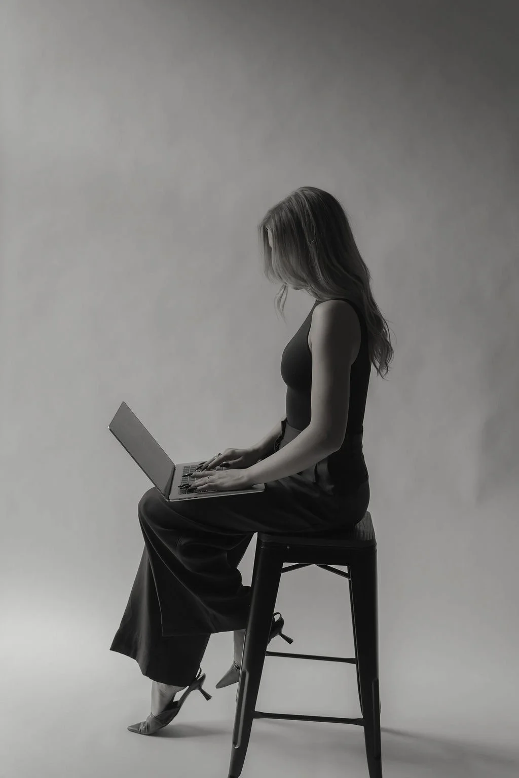 A woman sitting on a high stool working on a laptop, wearing a dark sleeveless top and wide-leg pants, in a monochrome setting.