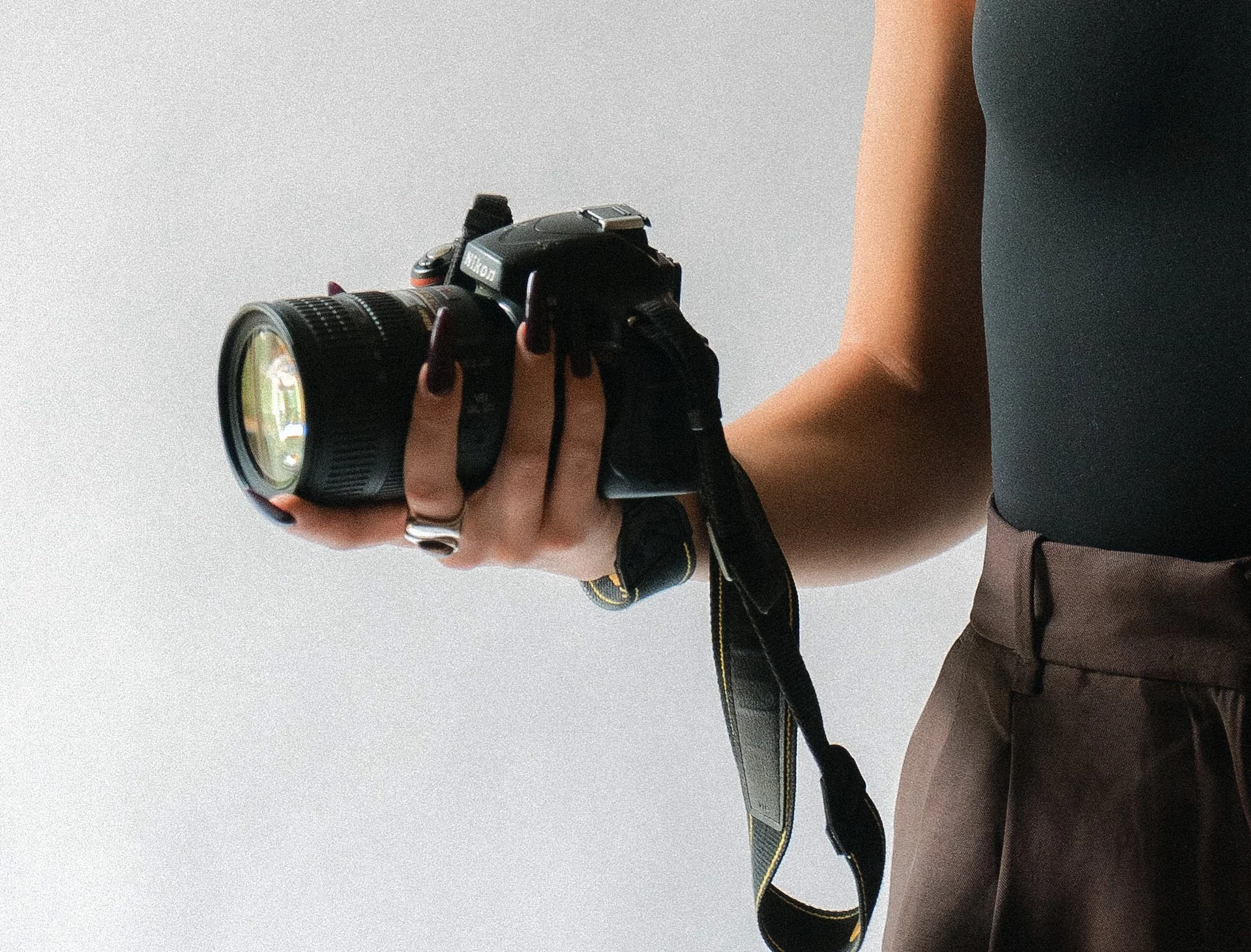 Person holding a Nikon DSLR camera pointing downward, wearing dark top and brown pants, with a plain light-colored wall background.