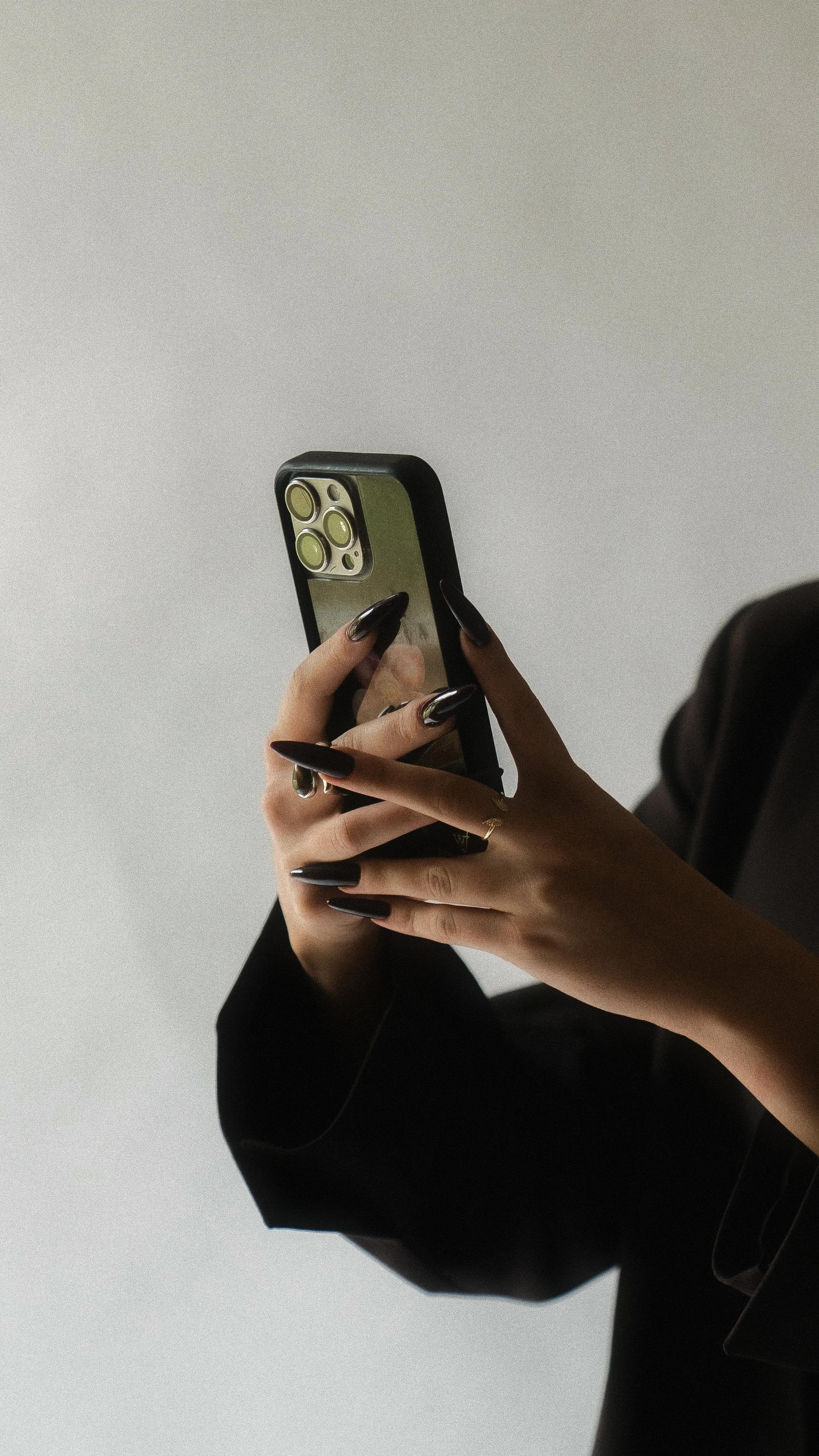 Close-up of a person's hands holding a smartphone with a reflective case, wearing dark long nails and a black outfit.