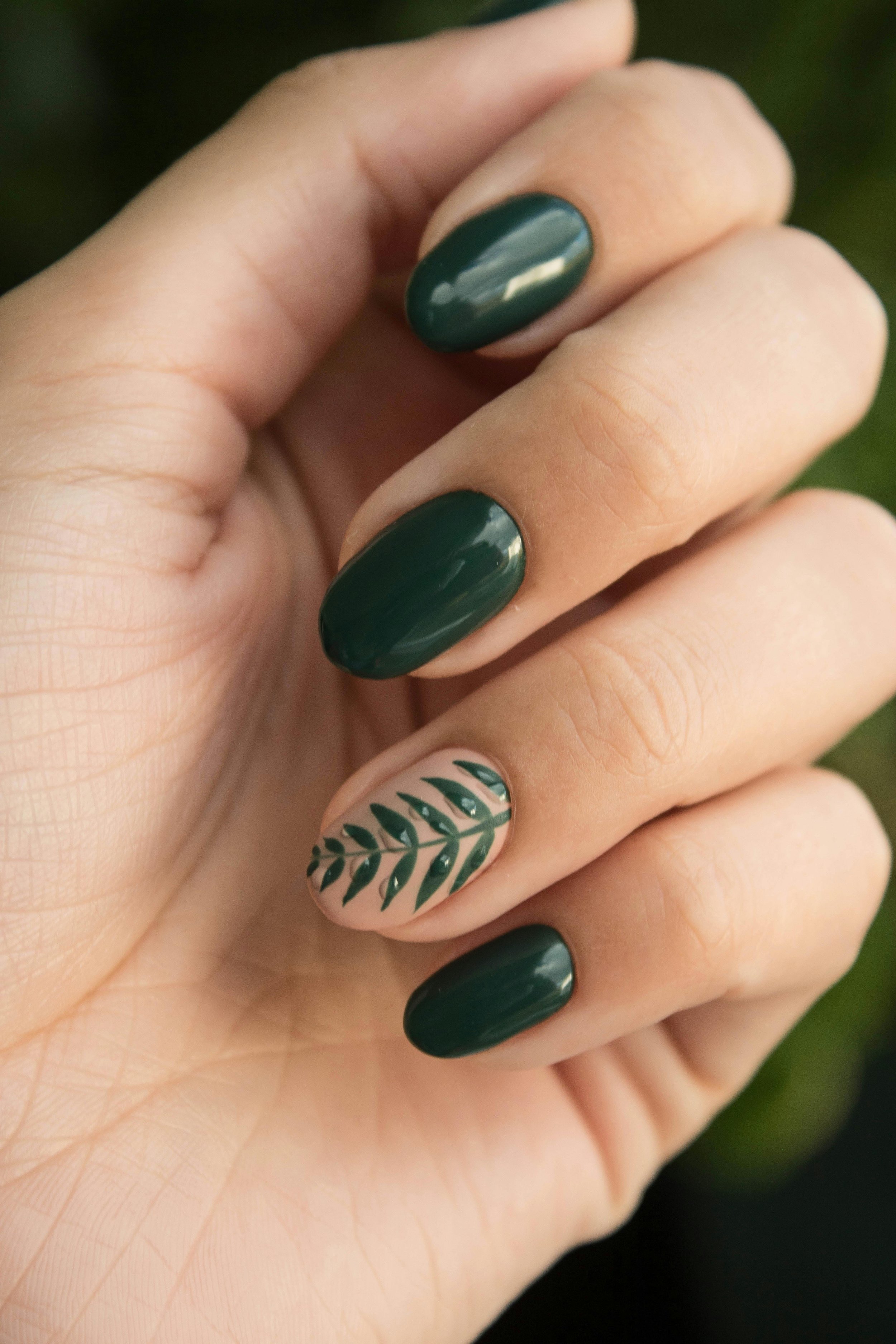 Close-up of a hand showing manicured nails painted in dark green, with one nail featuring a beige base with green leaf design and small rhinestones.