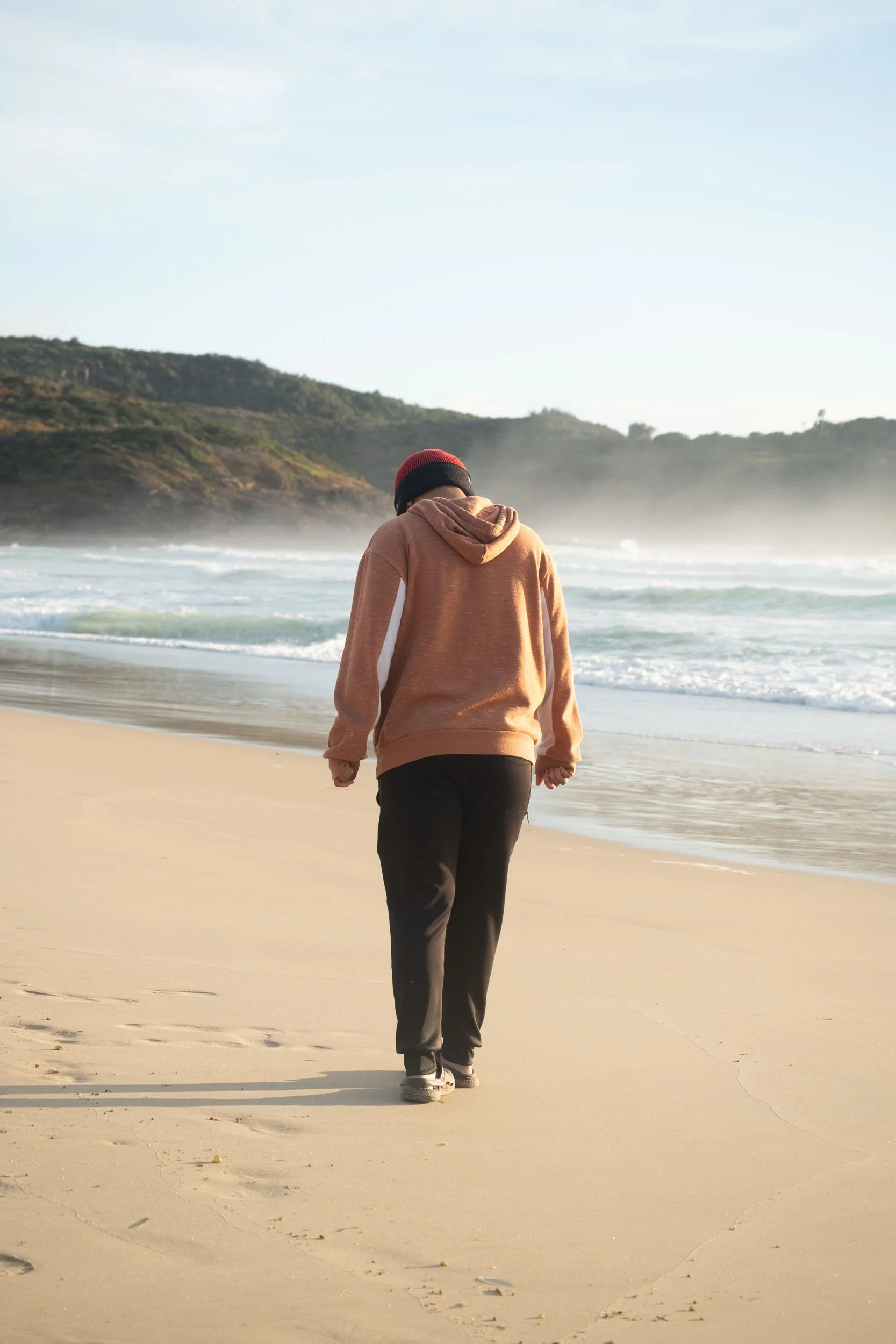 Person walking alone on the beach during sunrise or sunset, wearing a hoodie, black pants, and sneakers.