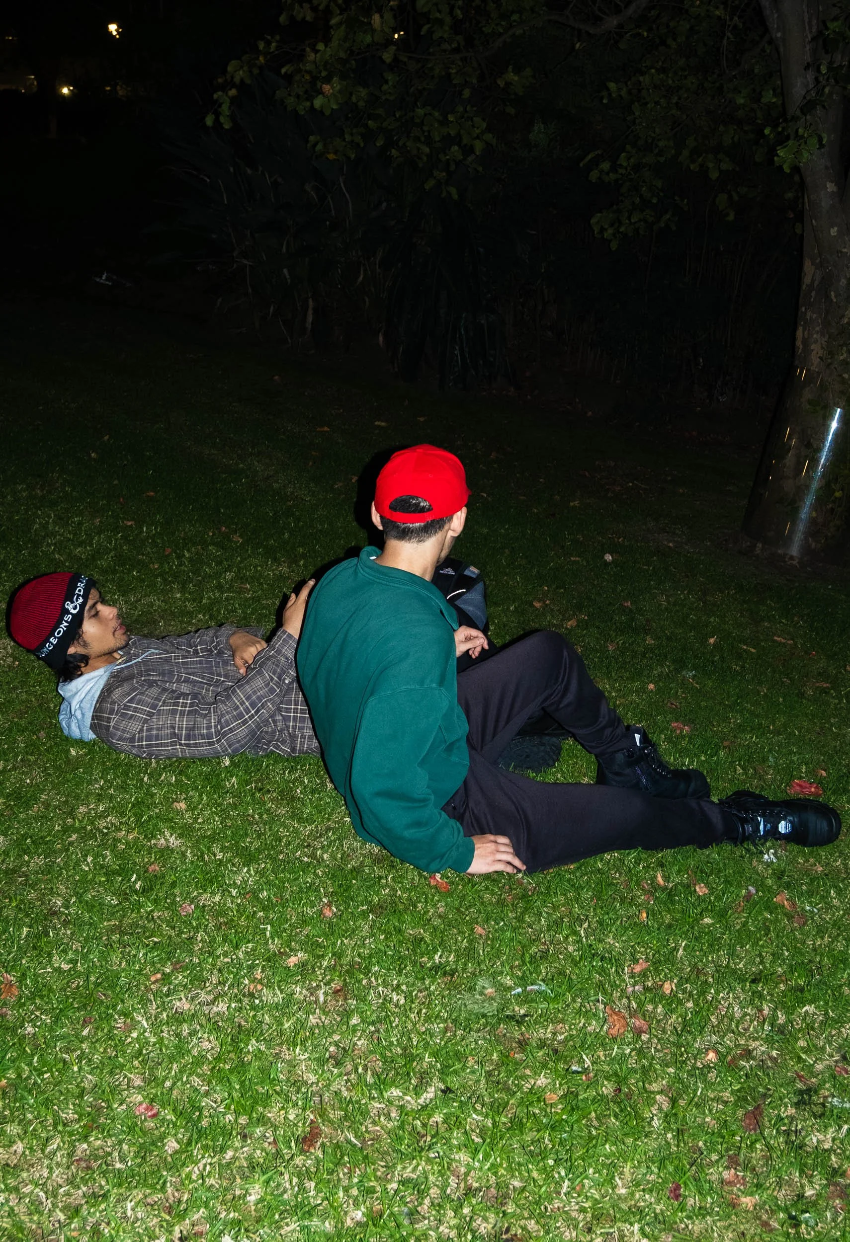 Two young men sitting on grass at night, one lying down and the other sitting with legs stretched out, near trees and a reflective surface.