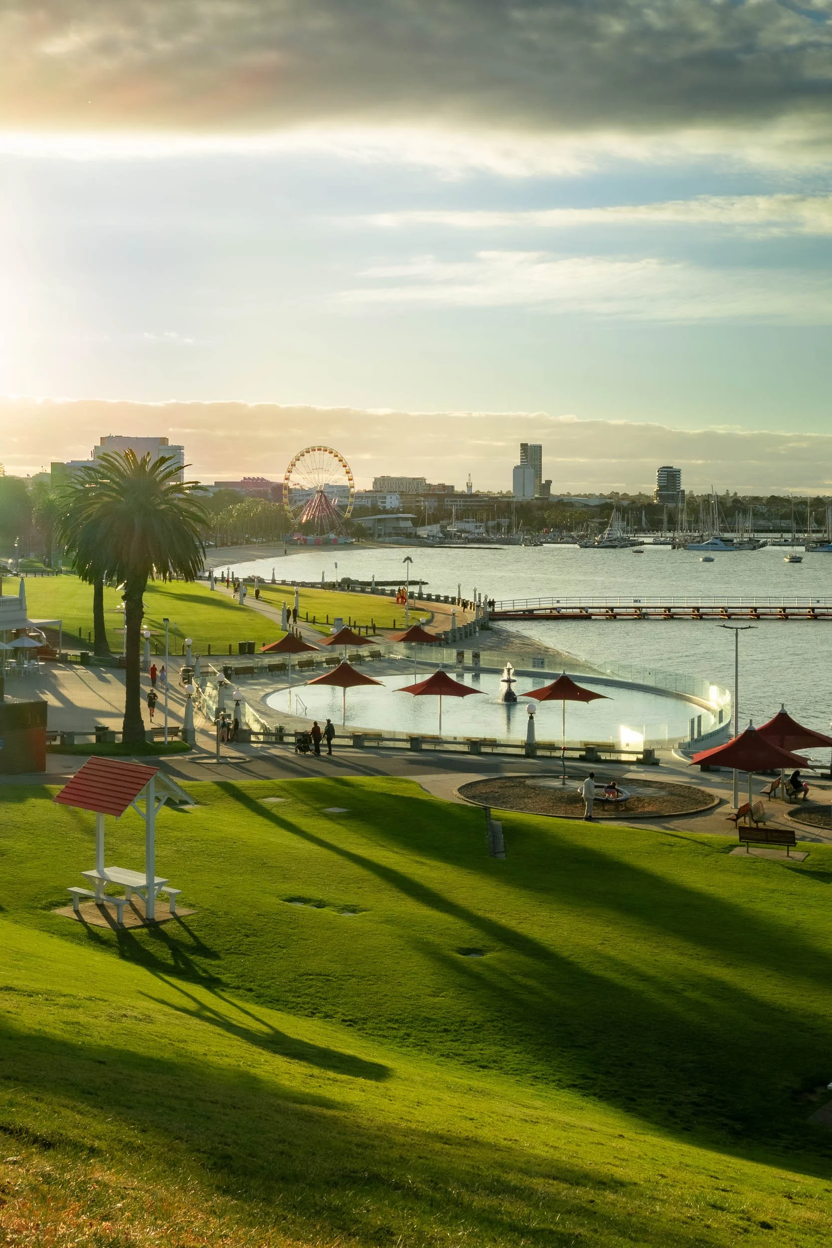 A scenic view of a park near water with green grass, trees, a fountain, and a pier, featuring a city skyline in the distance with a ferris wheel and boats on the water, under a partly cloudy sky during sunset.