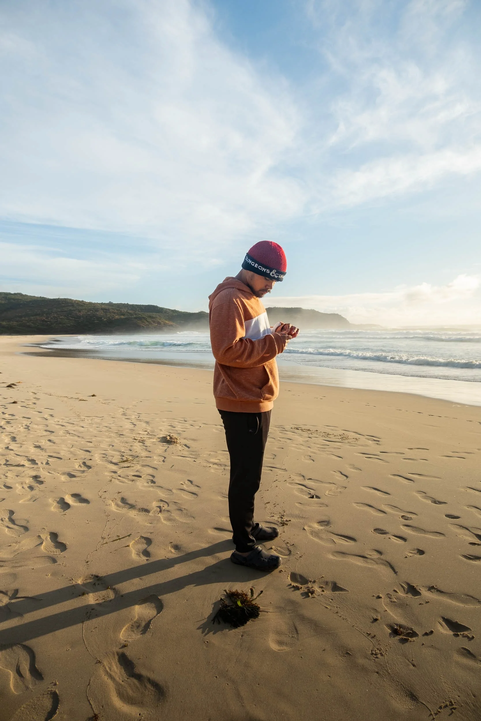 Person in a brown hoodie and black pants standing on a sandy beach, looking at their phone, with the ocean and cloudy sky in the background.