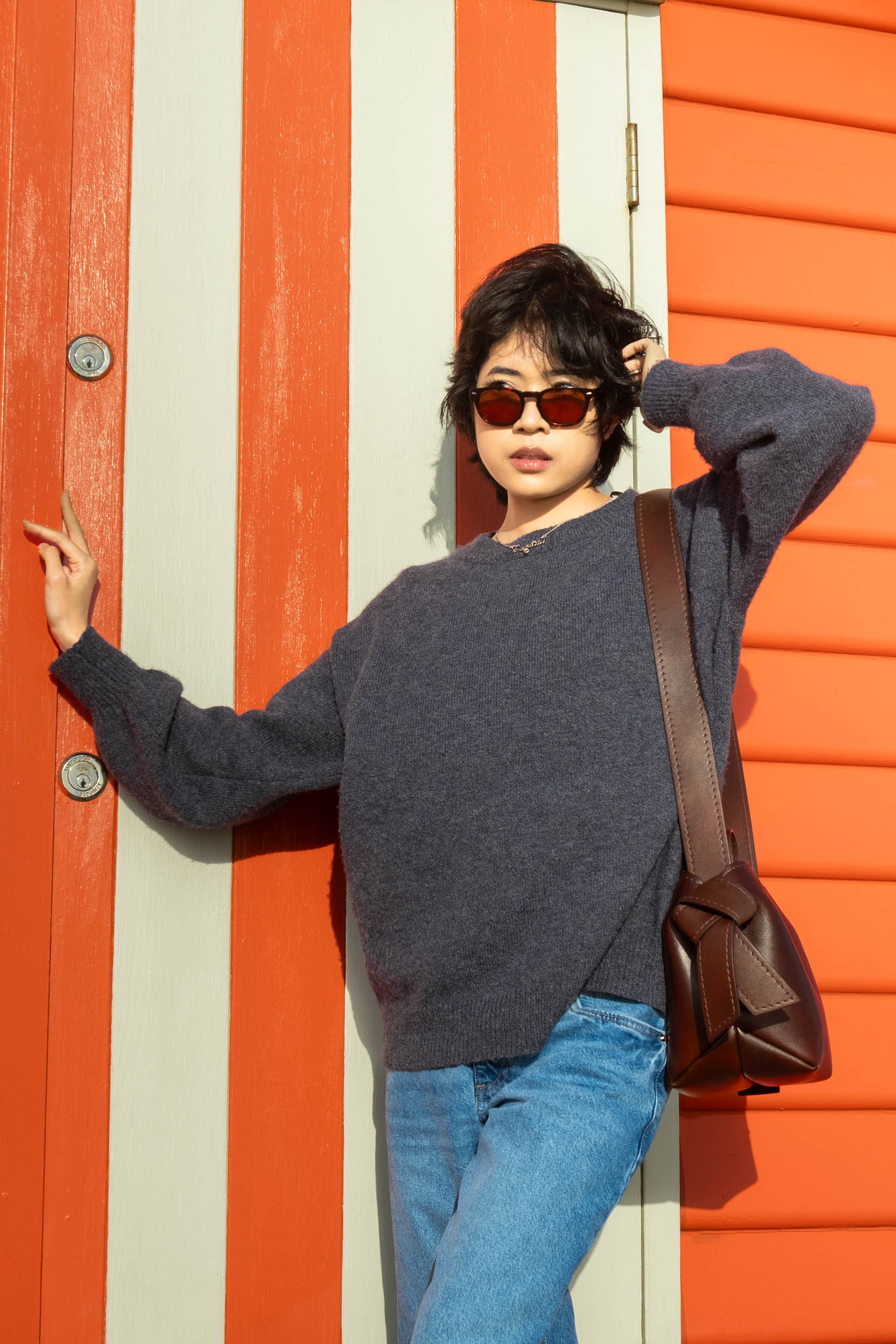 A woman with short dark hair wearing sunglasses, a dark gray oversized sweater, and blue jeans, standing against a colorful striped wall with orange and cream panels, holding her left hand near her head and touching the wall with her right hand.
