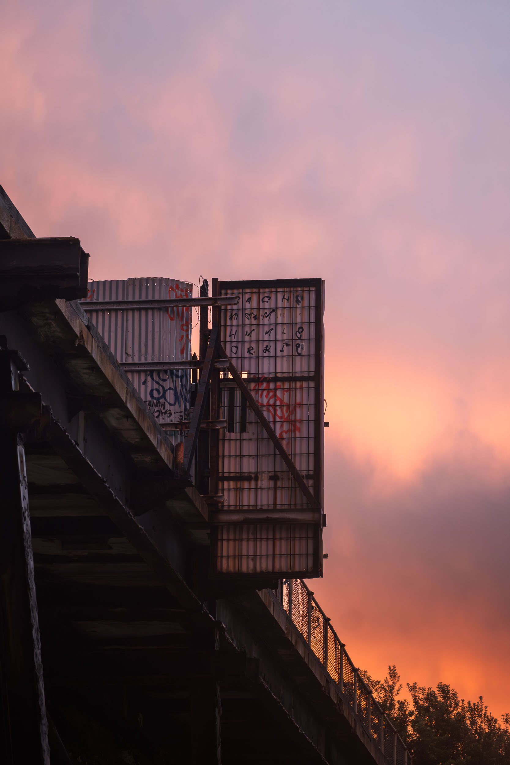 An urban scene showing the underside of a bridge with a fence and a billboard above, against a colorful sunset sky with pink and purple clouds.