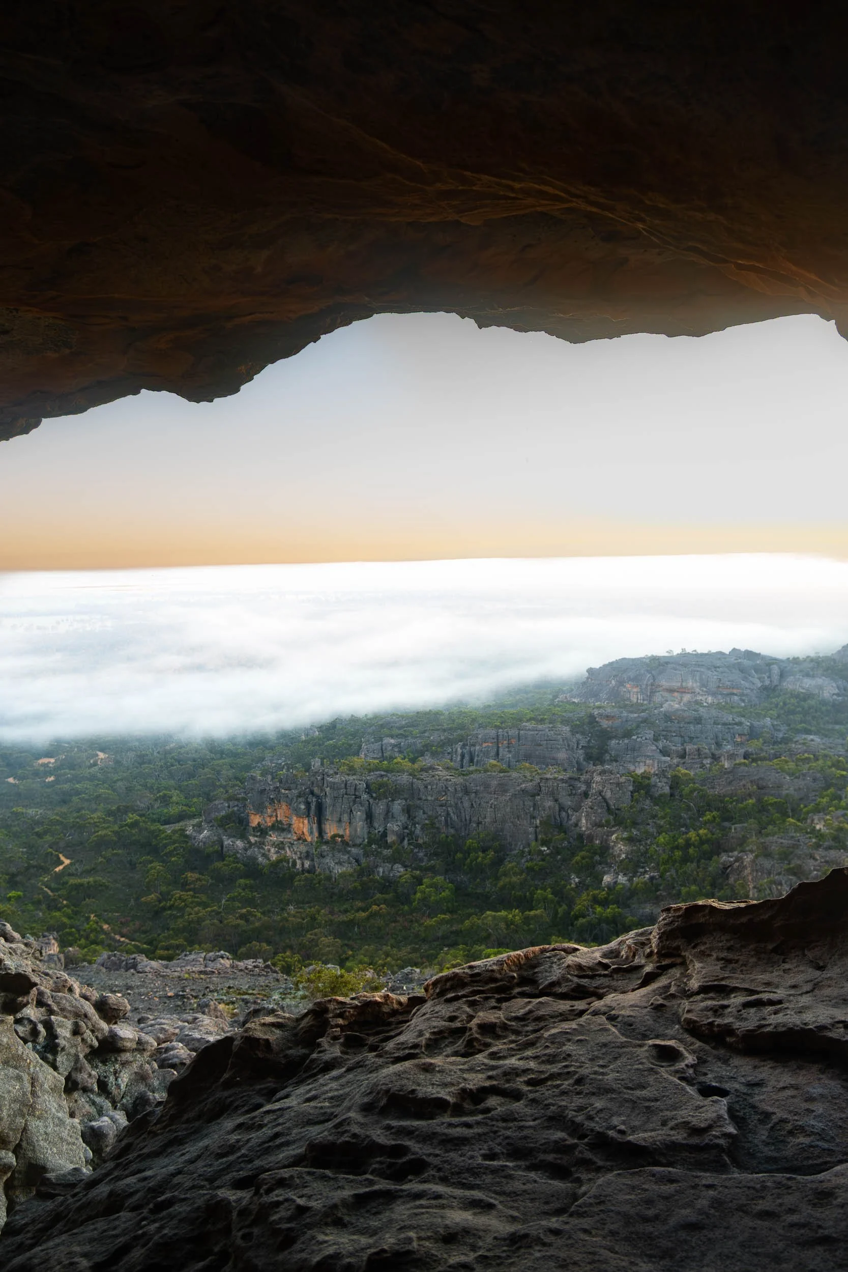 View of rocky landscape and forest seen from inside a cave with an opening, overlooking a foggy valley during sunrise or sunset.