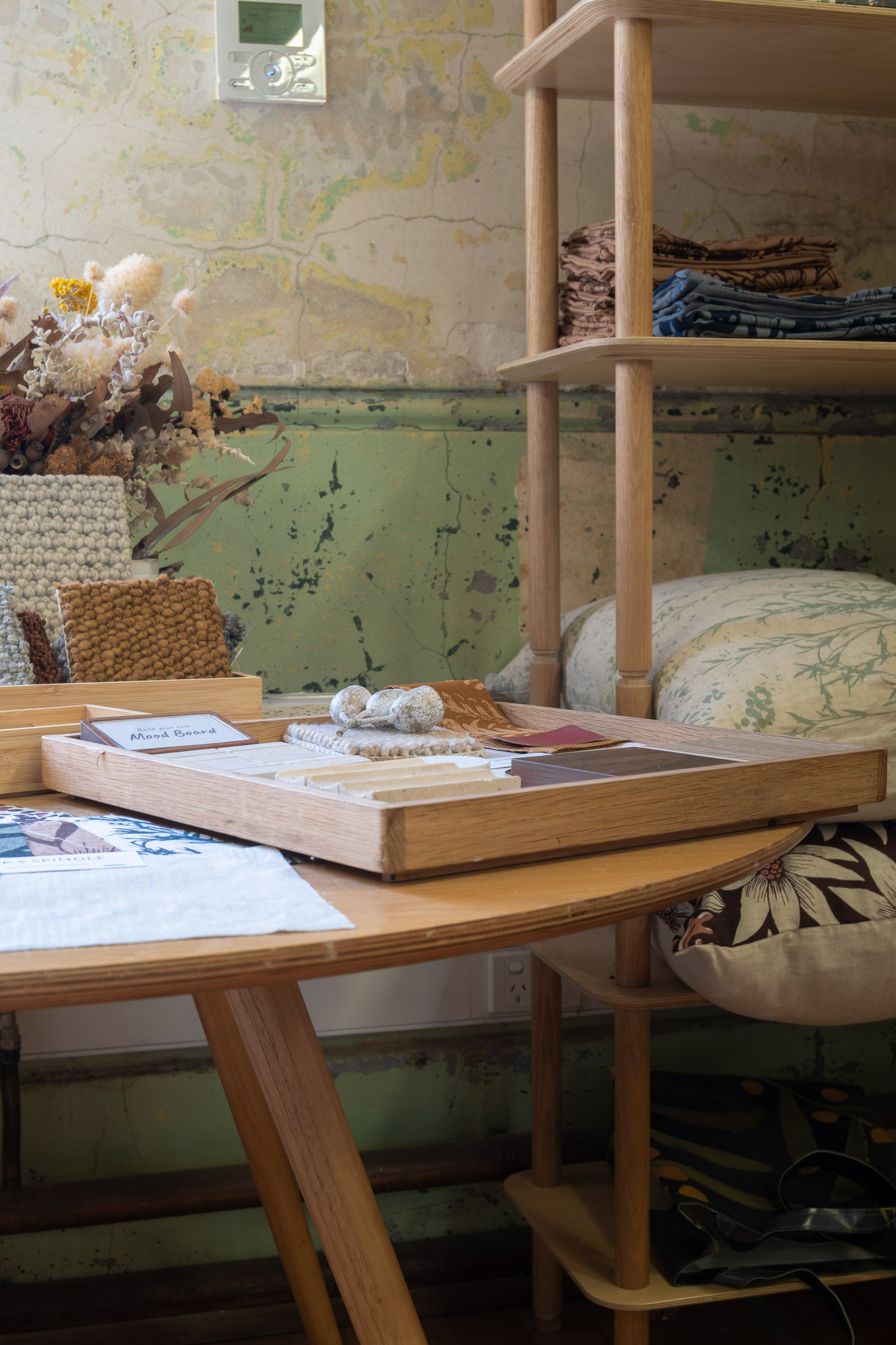 A cozy interior corner with a wooden table holding decorative items and a tray, a floral pillow, a bed with floral bedding, open shelves with folded clothes, a textured wall, and dried flowers in a vase.