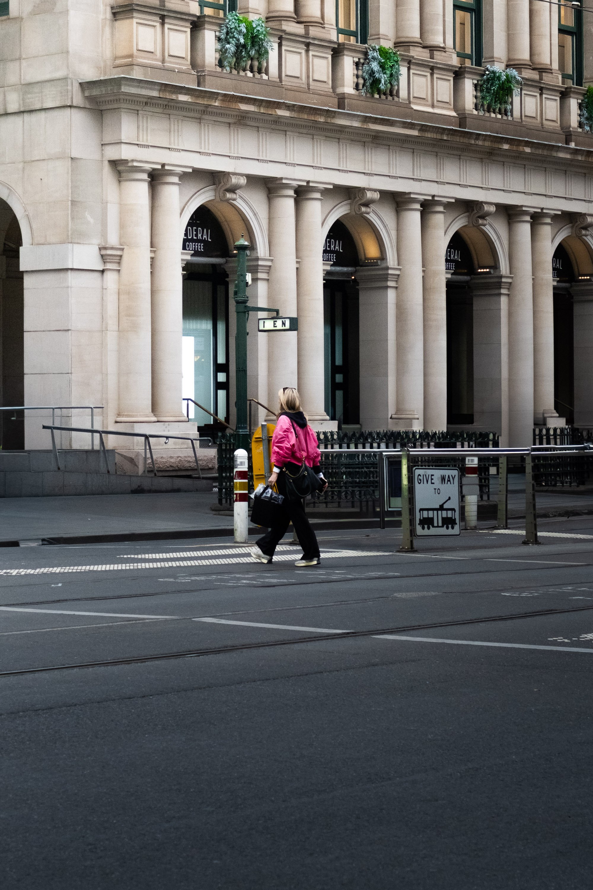 A woman in a pink jacket and black pants walking across a street in front of a historic building with large columns and decorative planters. She is carrying a black bag and a suitcase.